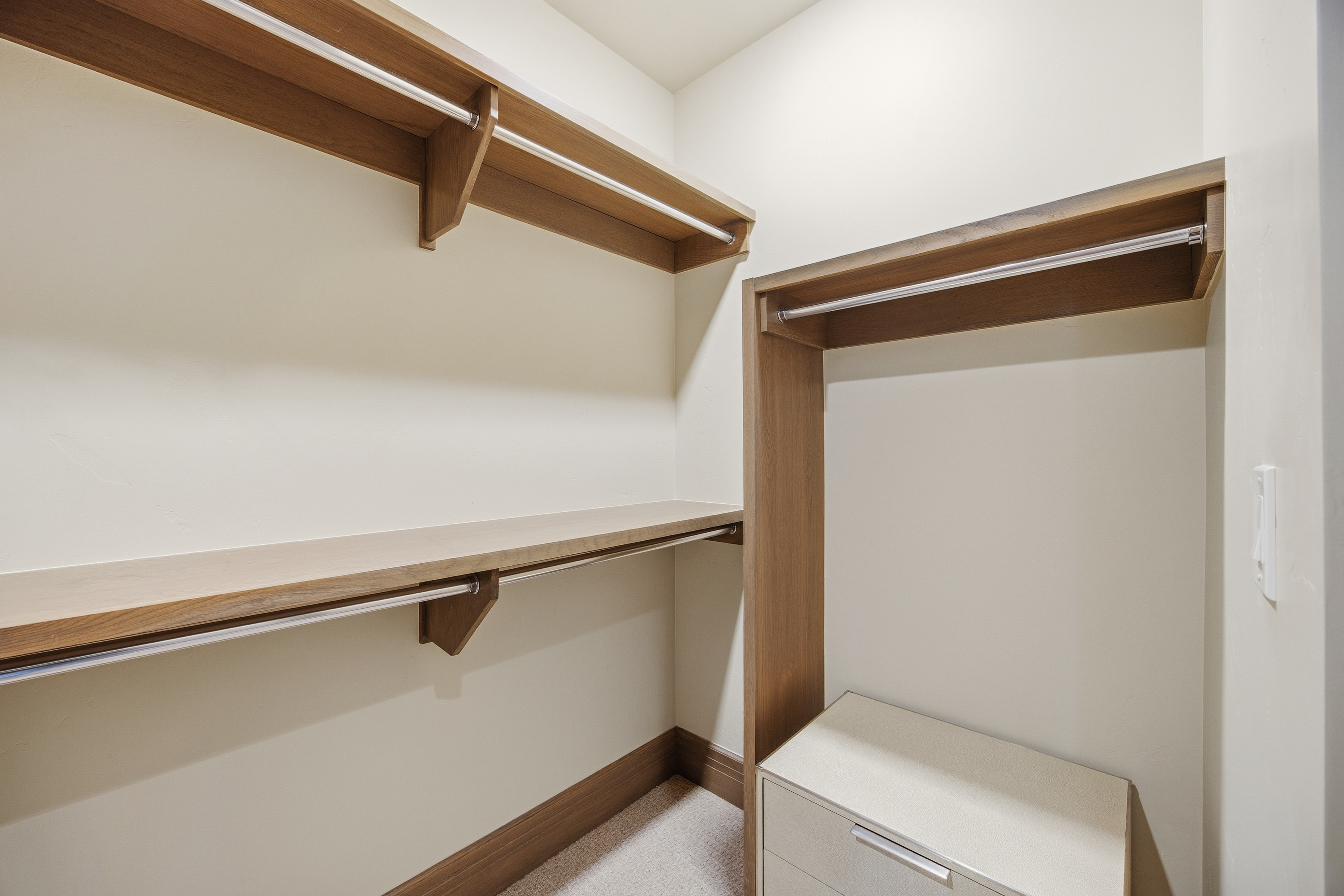 This is a well-organized walk-in closet featuring wooden shelving and hanging rods. The closet has a neutral color scheme with light beige walls and wood trim, creating a clean and functional space. A small dresser is visible in the corner, adding to the storage options.