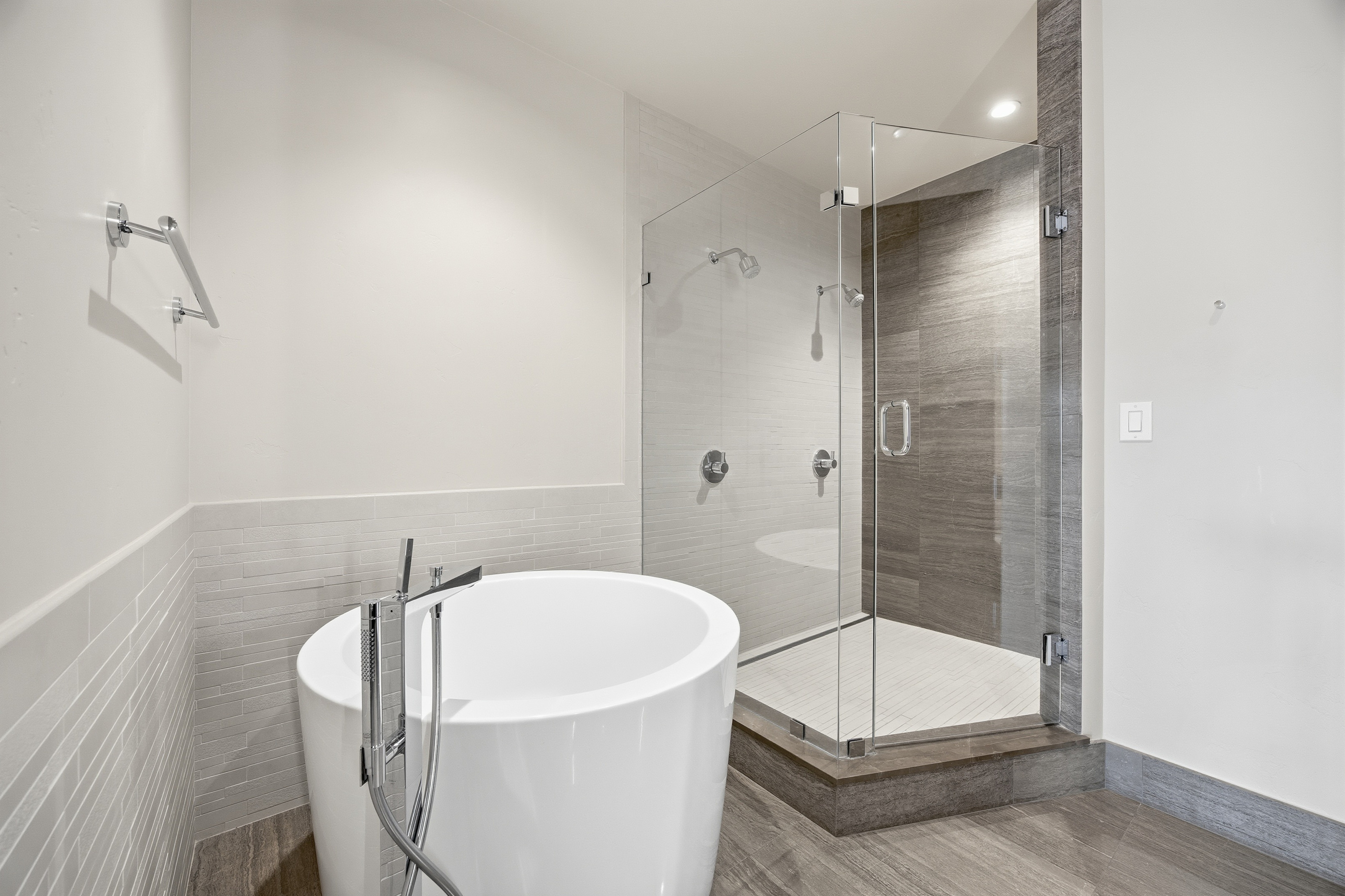 This is a bright and modern primary bathroom featuring a freestanding white bathtub with a chrome faucet, a glass-enclosed shower with dual shower heads, and neutral-toned tiling. The flooring is a wood-look tile, and the walls are painted a clean white, creating a spa-like atmosphere. The perspective is a wide shot, showcasing the layout and key features of the bathroom.