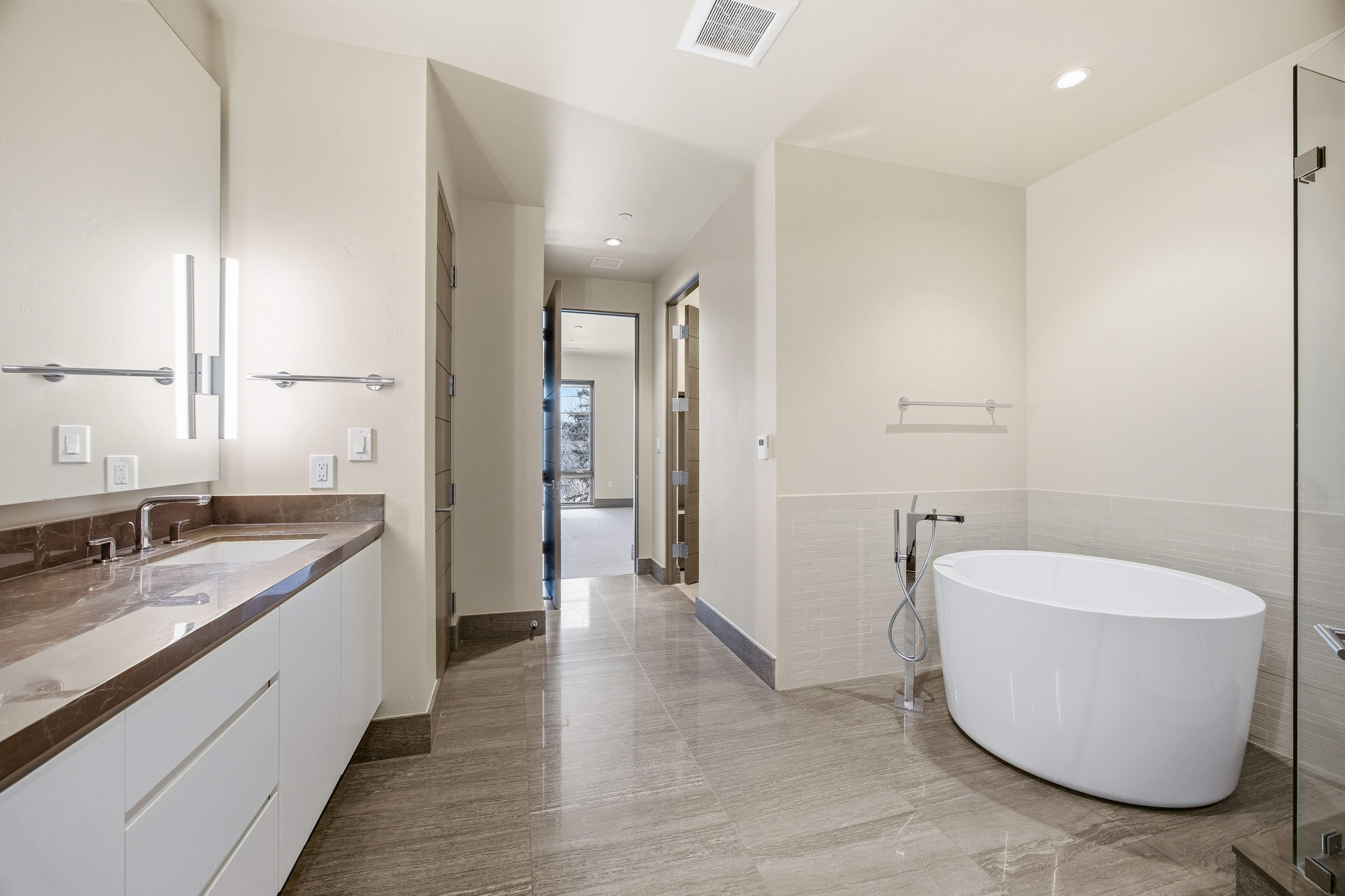 This is a bright and modern primary bathroom featuring a freestanding white bathtub, a sleek vanity with a brown countertop, and contemporary fixtures. The bathroom has neutral-toned walls and tiled flooring, creating a clean and luxurious atmosphere. The perspective is from the doorway, showcasing the spacious layout and high-end finishes.