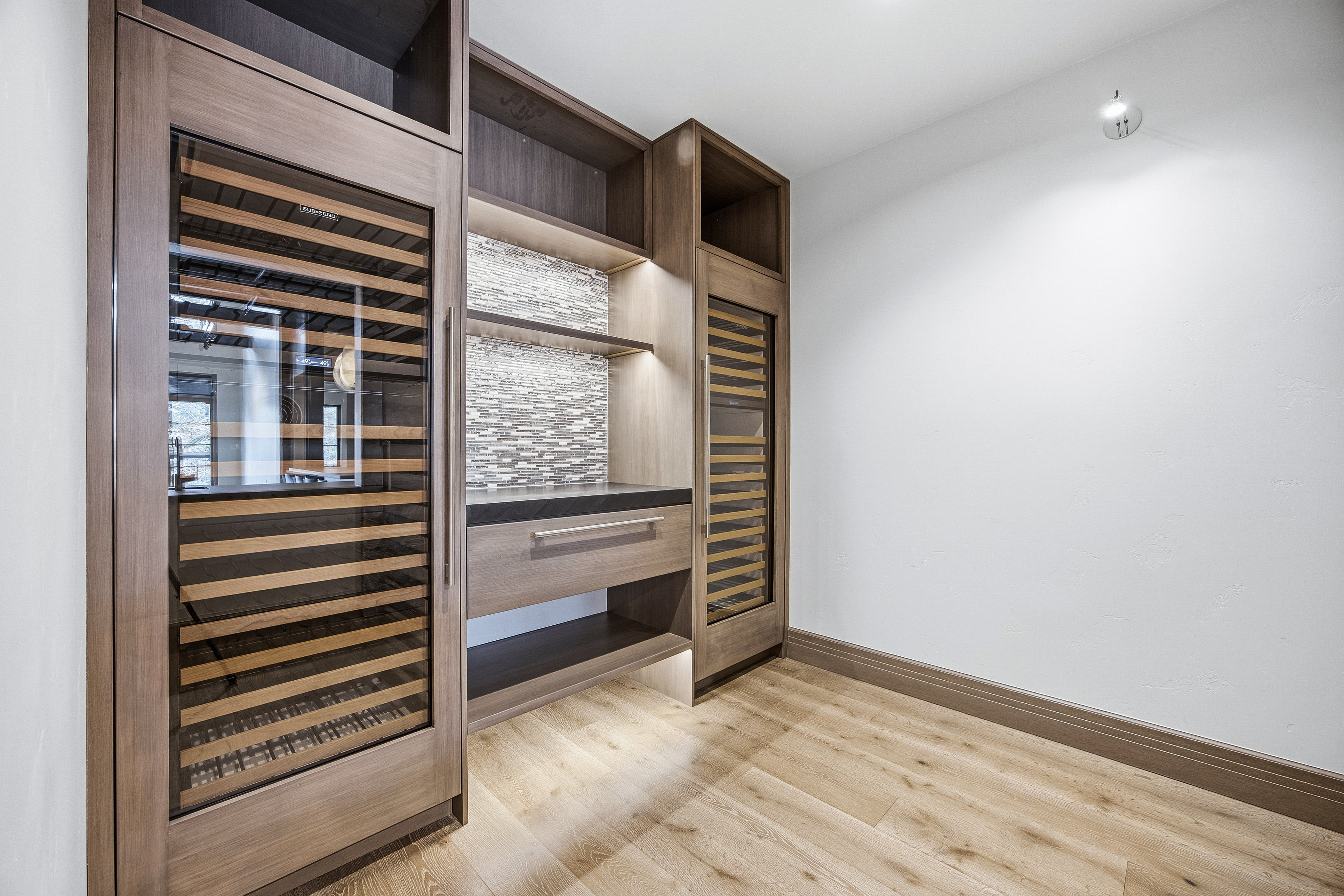 This interior shot showcases a modern wine cellar with built-in wooden wine racks and a sleek countertop area. The design incorporates a mosaic tile backsplash and recessed lighting, creating an elegant and functional space for wine storage and display. The light wood flooring adds warmth to the room.