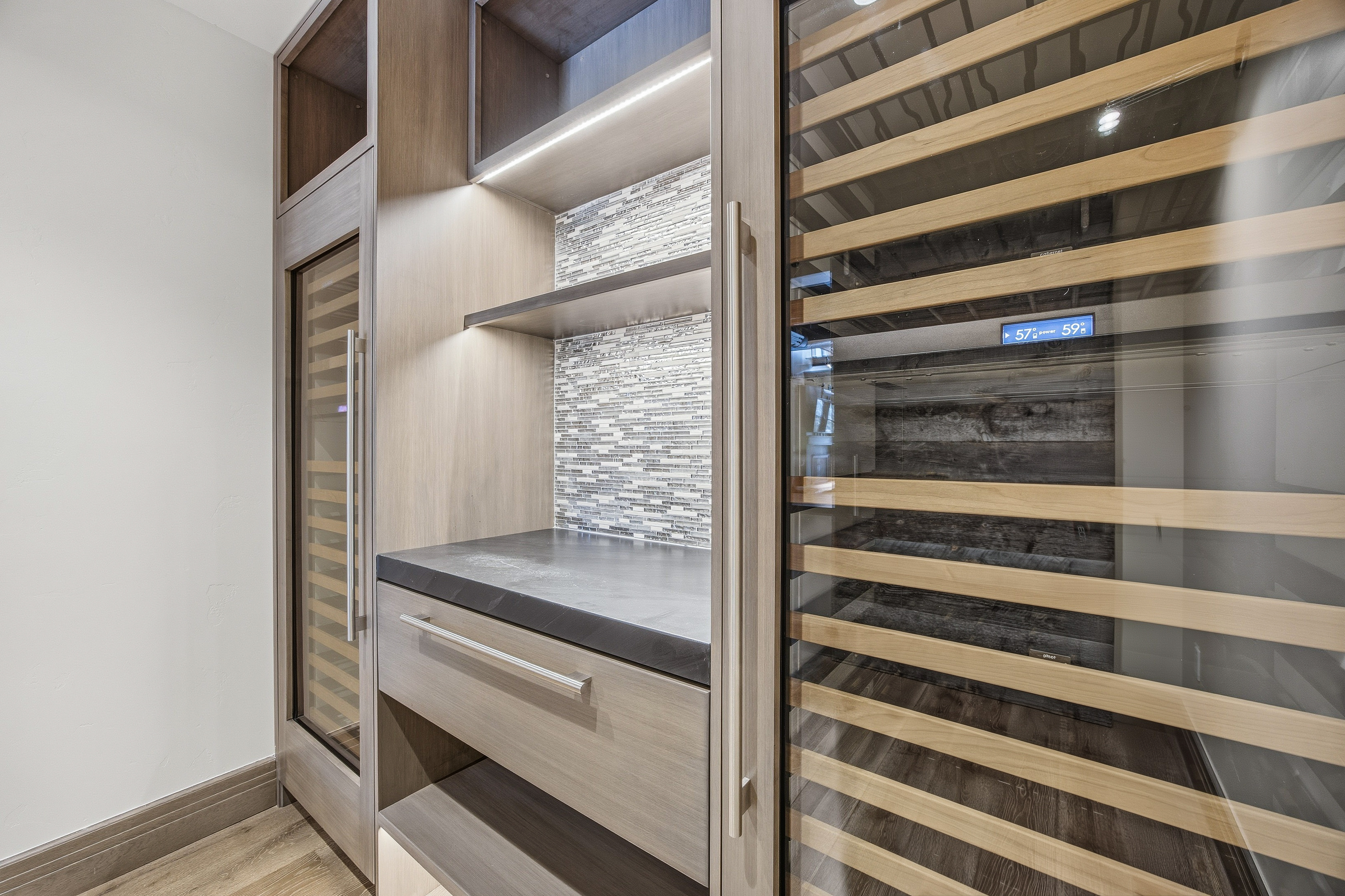 This image showcases a modern wine cellar, featuring a built-in wine refrigerator with wooden racks and a digital temperature display. Adjacent to the refrigerator is a storage area with shelves, a countertop, and a drawer, accented by a mosaic tile backsplash and integrated lighting. The overall design is sleek and sophisticated, emphasizing both functionality and aesthetic appeal.