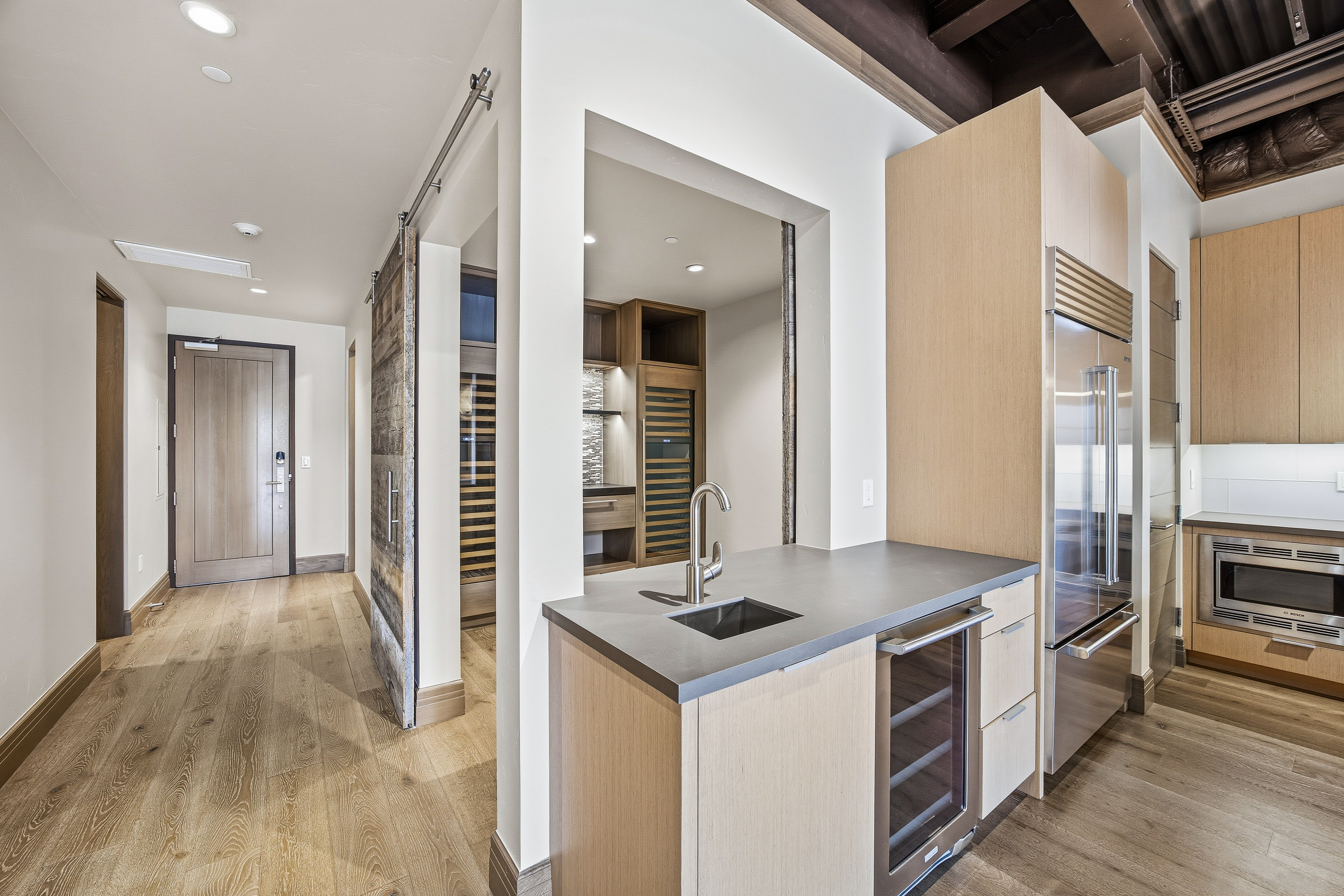 This interior shot showcases a modern kitchen area with light wood cabinetry, stainless steel appliances, and a gray countertop. A wine fridge is integrated into the cabinetry, and a rustic sliding barn door adds a touch of character. The space is well-lit and appears to be part of a larger open-concept design.