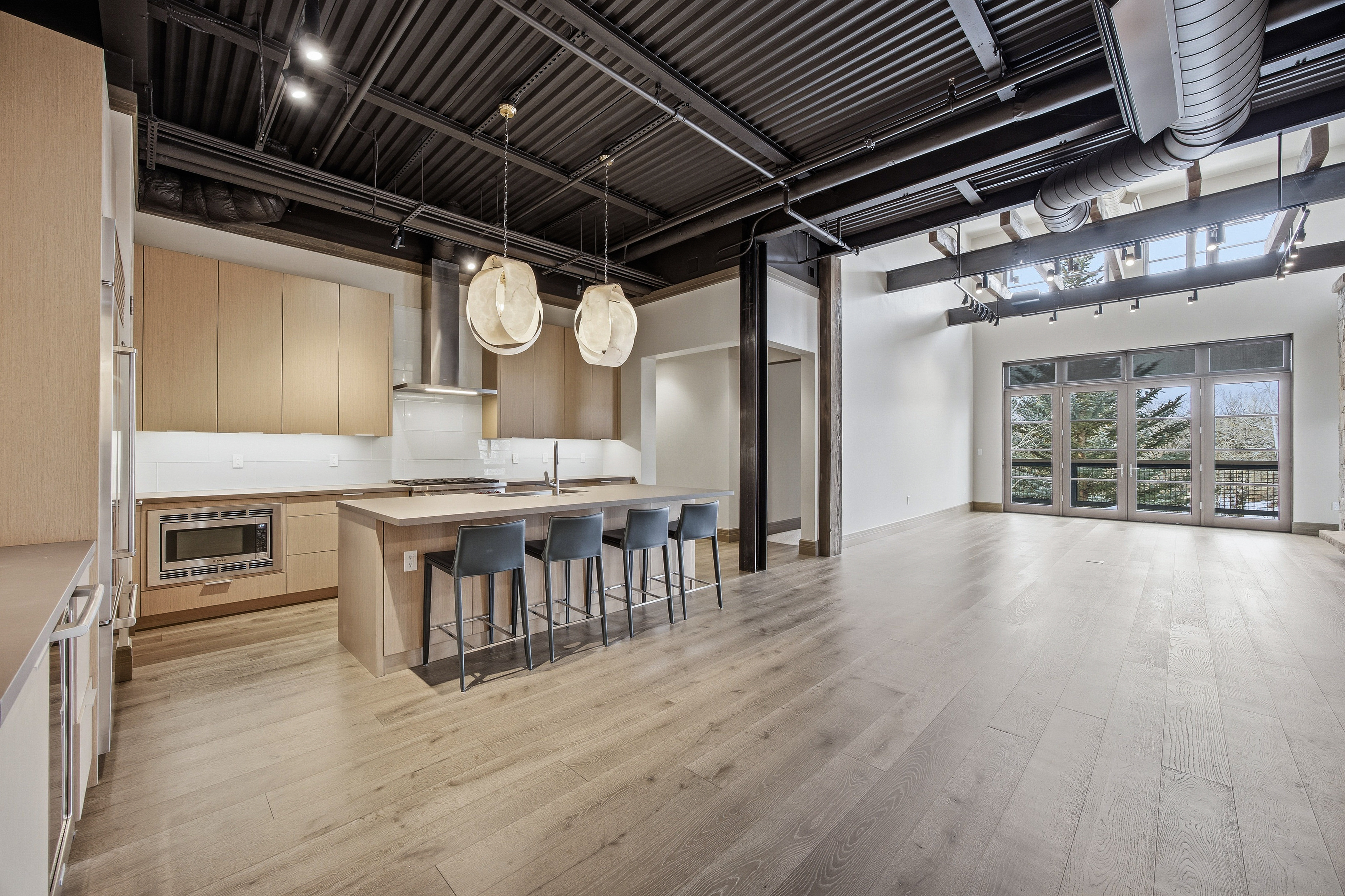 This interior shot showcases a modern kitchen and living area with an open floor plan. The kitchen features light wood cabinetry, stainless steel appliances, and a central island with bar seating, illuminated by pendant lights. The living area boasts high ceilings with exposed ductwork and large windows offering natural light, creating a spacious and contemporary atmosphere.