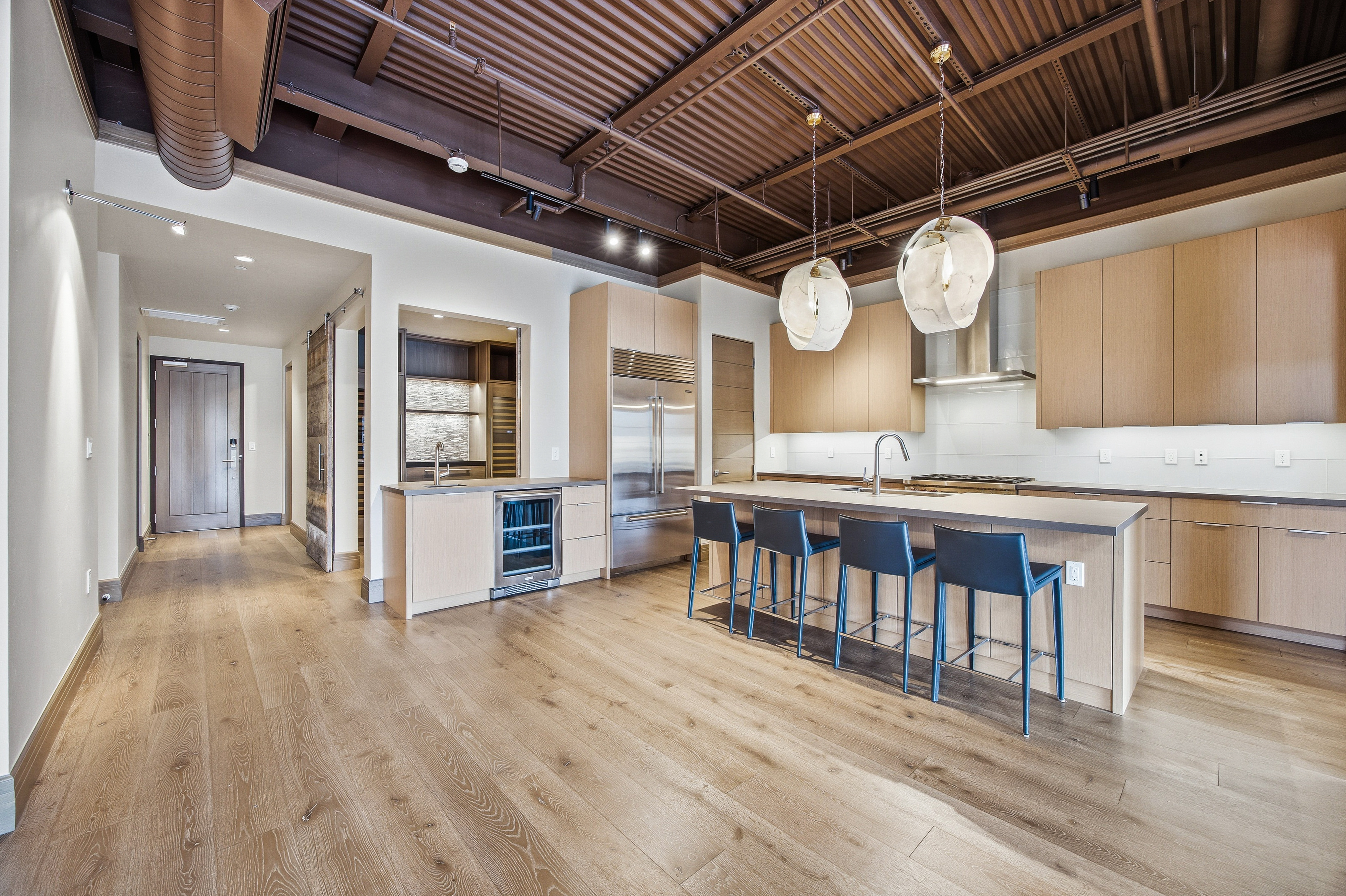 This is a spacious, modern kitchen featuring light wood cabinetry, stainless steel appliances, and a large island with seating. The open floor plan leads into a hallway with a sliding barn door, and exposed ductwork and pendant lighting add an industrial-chic touch. The overall impression is clean, contemporary, and inviting.