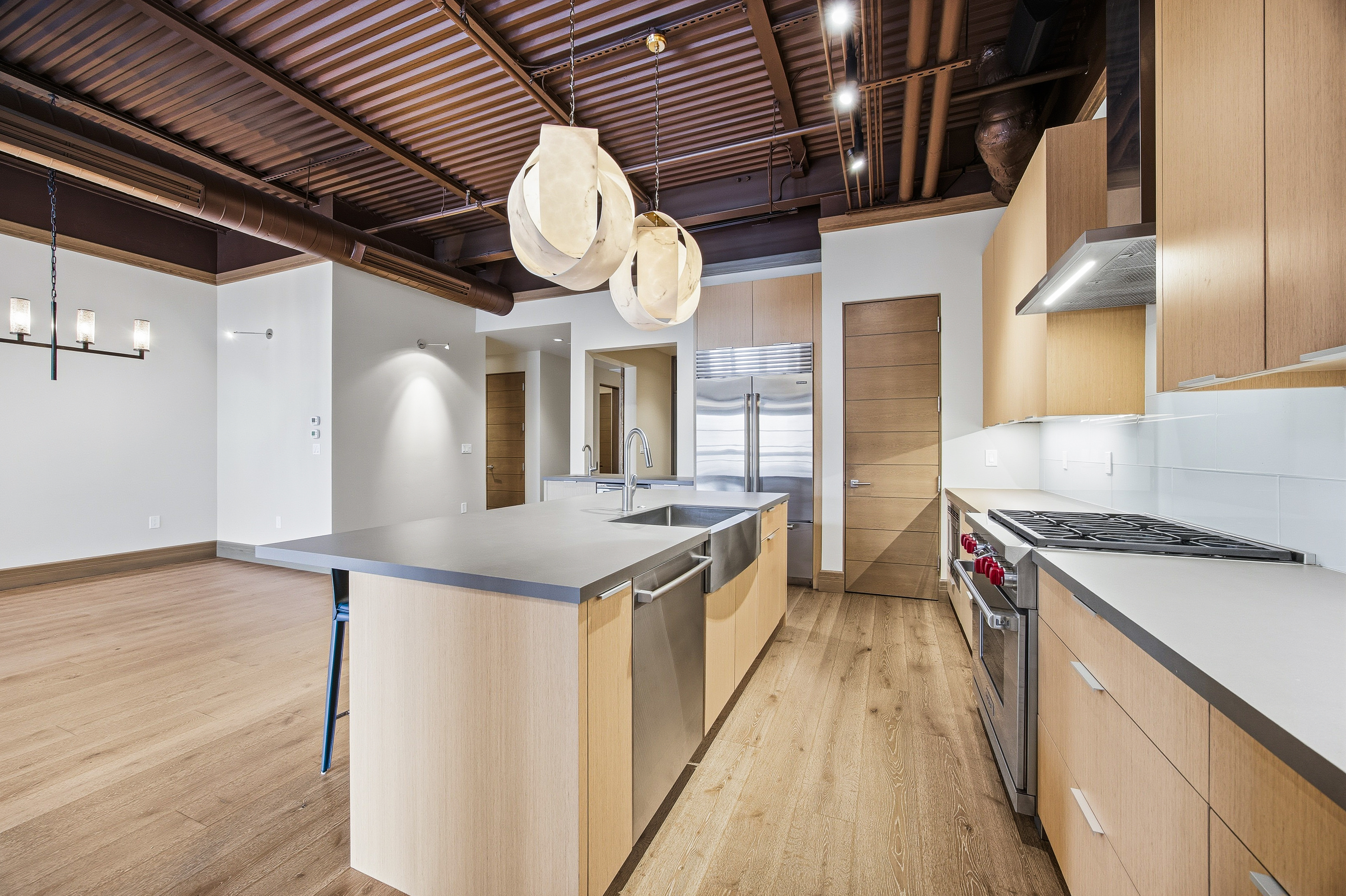 This is a modern kitchen with an open floor plan, featuring light wood cabinetry and stainless steel appliances. A large island with a gray countertop dominates the center of the room, illuminated by two unique pendant lights. The exposed ceiling adds an industrial touch, while the hardwood floors provide warmth and continuity throughout the space.