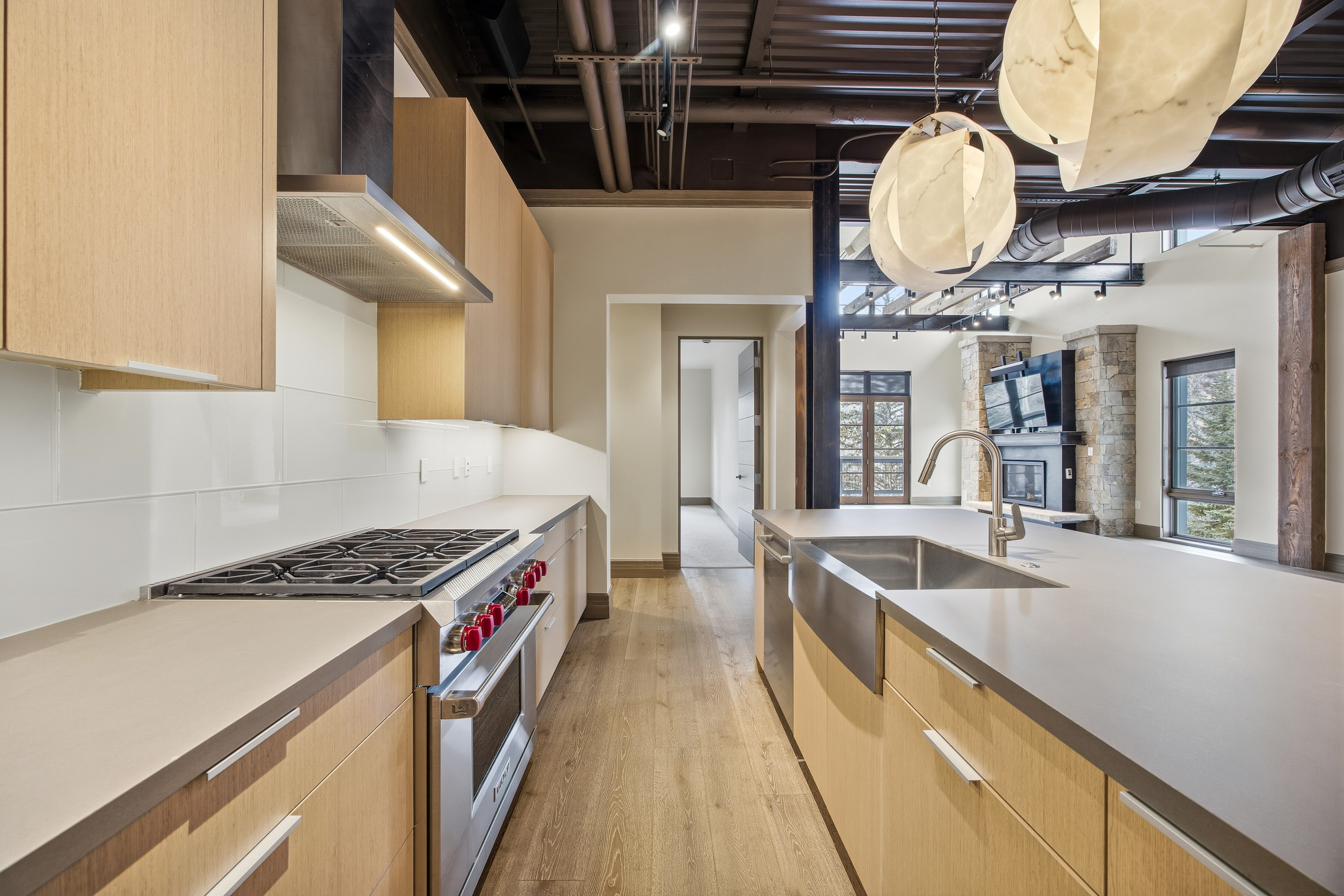 This is a well-lit, modern kitchen featuring light wood cabinetry, stainless steel appliances, and light countertops. The kitchen includes a gas range and a large island with a sink. The open floor plan leads into a living area with a stone fireplace, creating a seamless and inviting space.