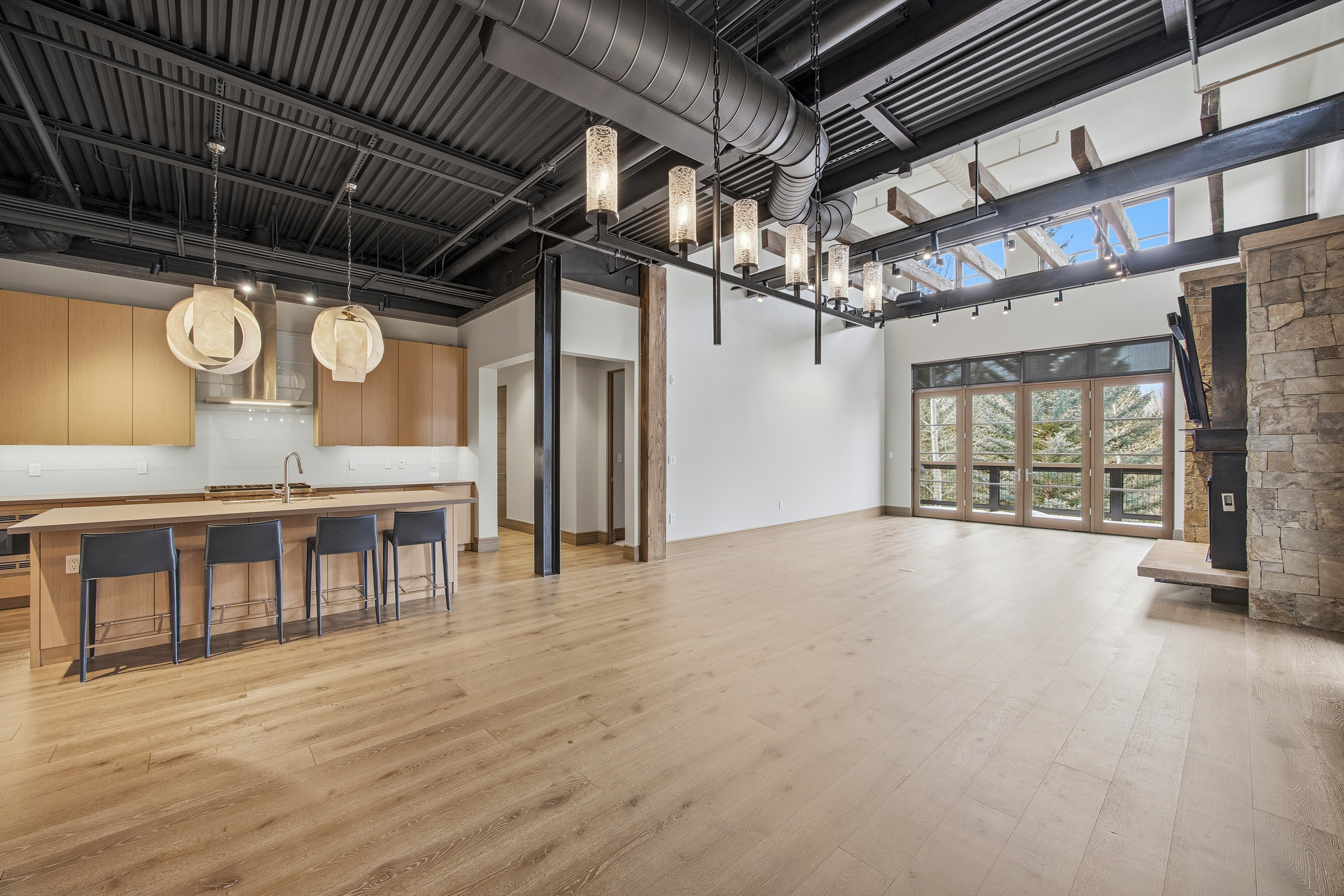 This is a spacious, modern living room featuring an open floor plan that integrates the kitchen area. The room boasts hardwood floors, a black exposed ceiling with industrial-style ductwork and lighting fixtures, and a stone fireplace. Large windows and doors provide ample natural light and views of the outdoors, creating a bright and airy atmosphere.