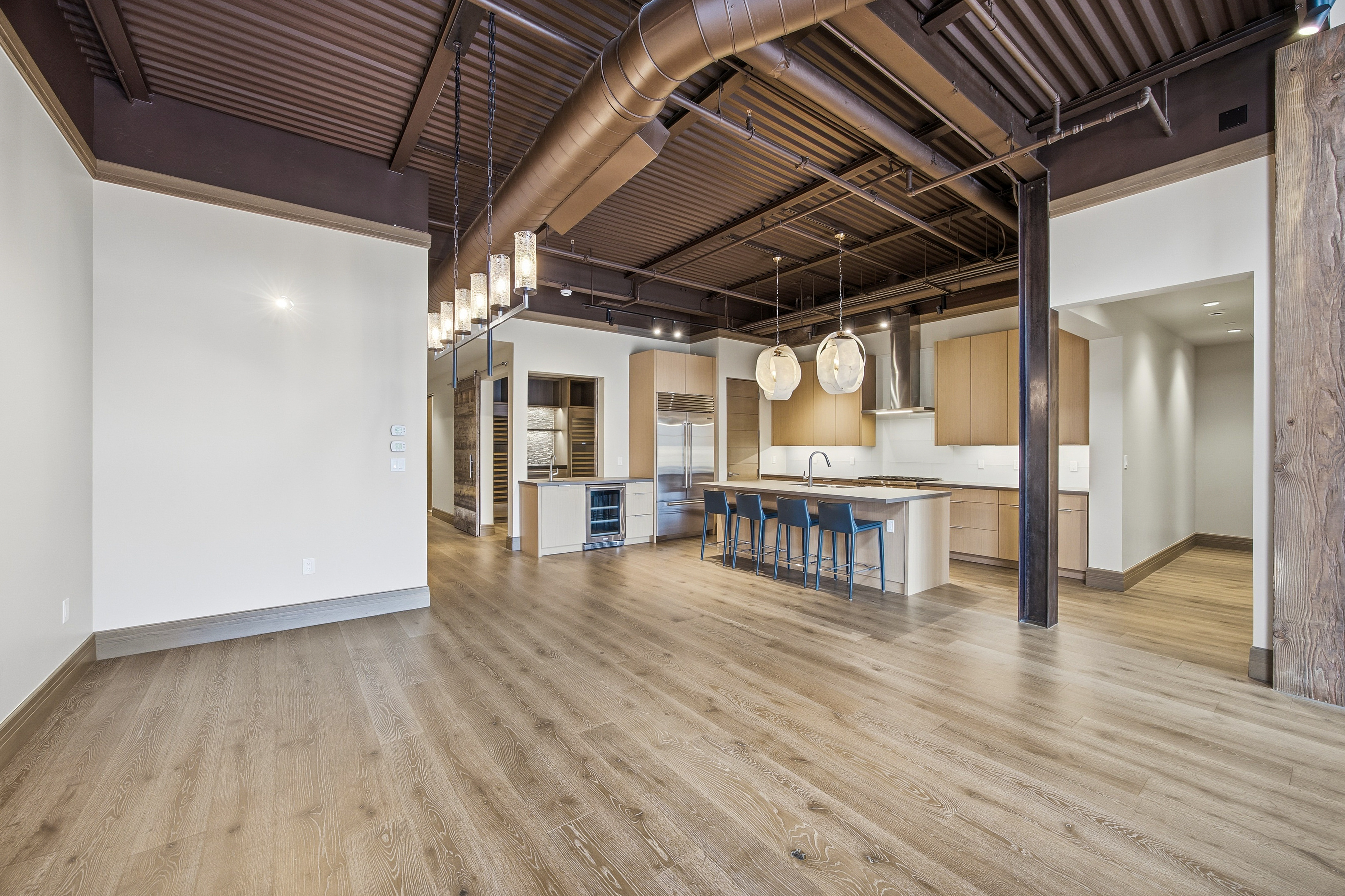 This is a wide-angle shot of a modern, open-concept kitchen and living area. The kitchen features light wood cabinetry, stainless steel appliances, and a large island with seating. Exposed ductwork and industrial-style lighting fixtures add an urban, loft-like feel to the space, while the light wood flooring provides warmth and continuity throughout.