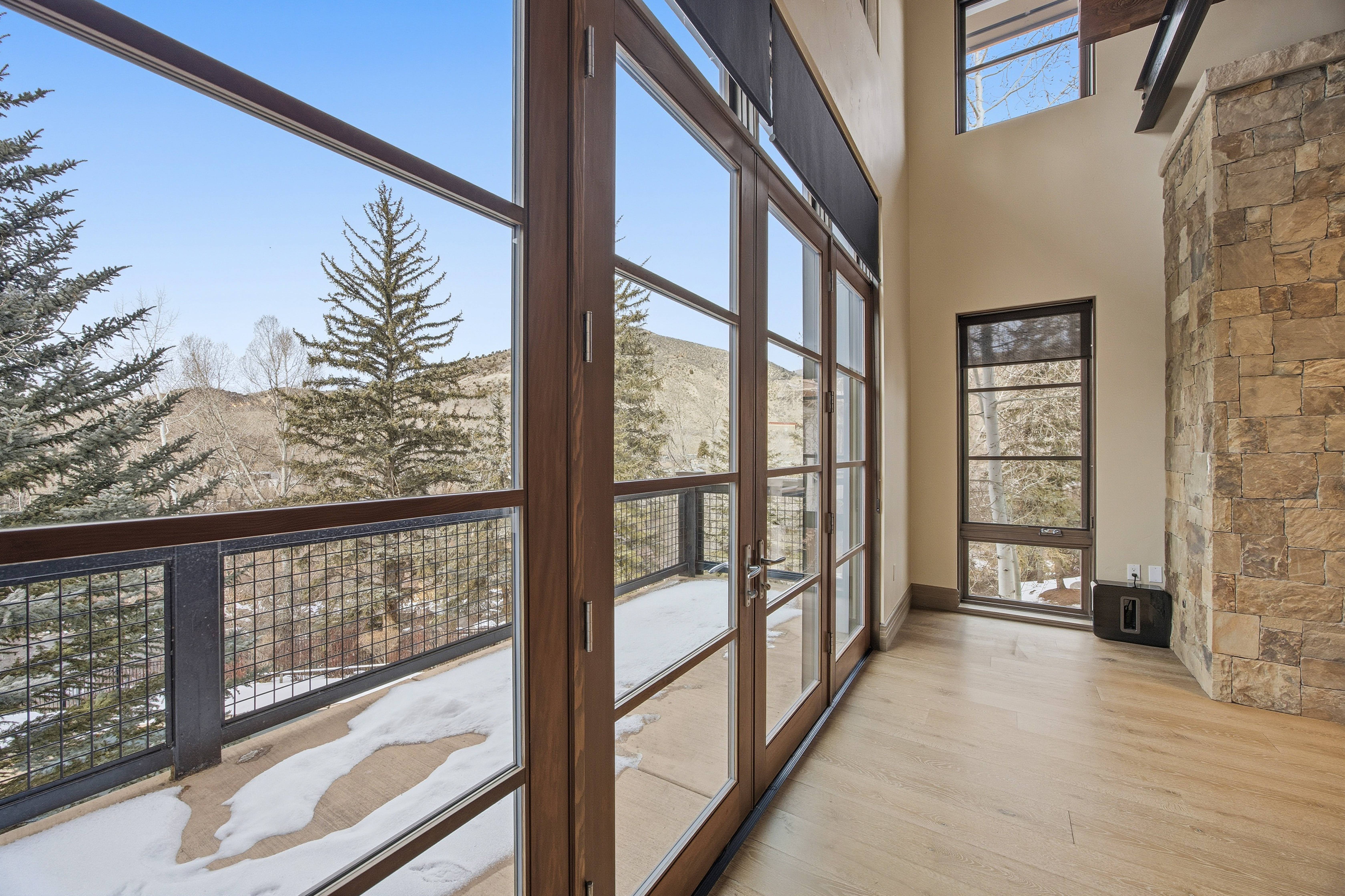 This interior shot showcases a bright living room with large windows offering scenic mountain views. The room features hardwood floors and a stone accent wall, creating a warm and inviting atmosphere. A balcony with a metal railing is accessible through the glass doors, blending indoor and outdoor living.