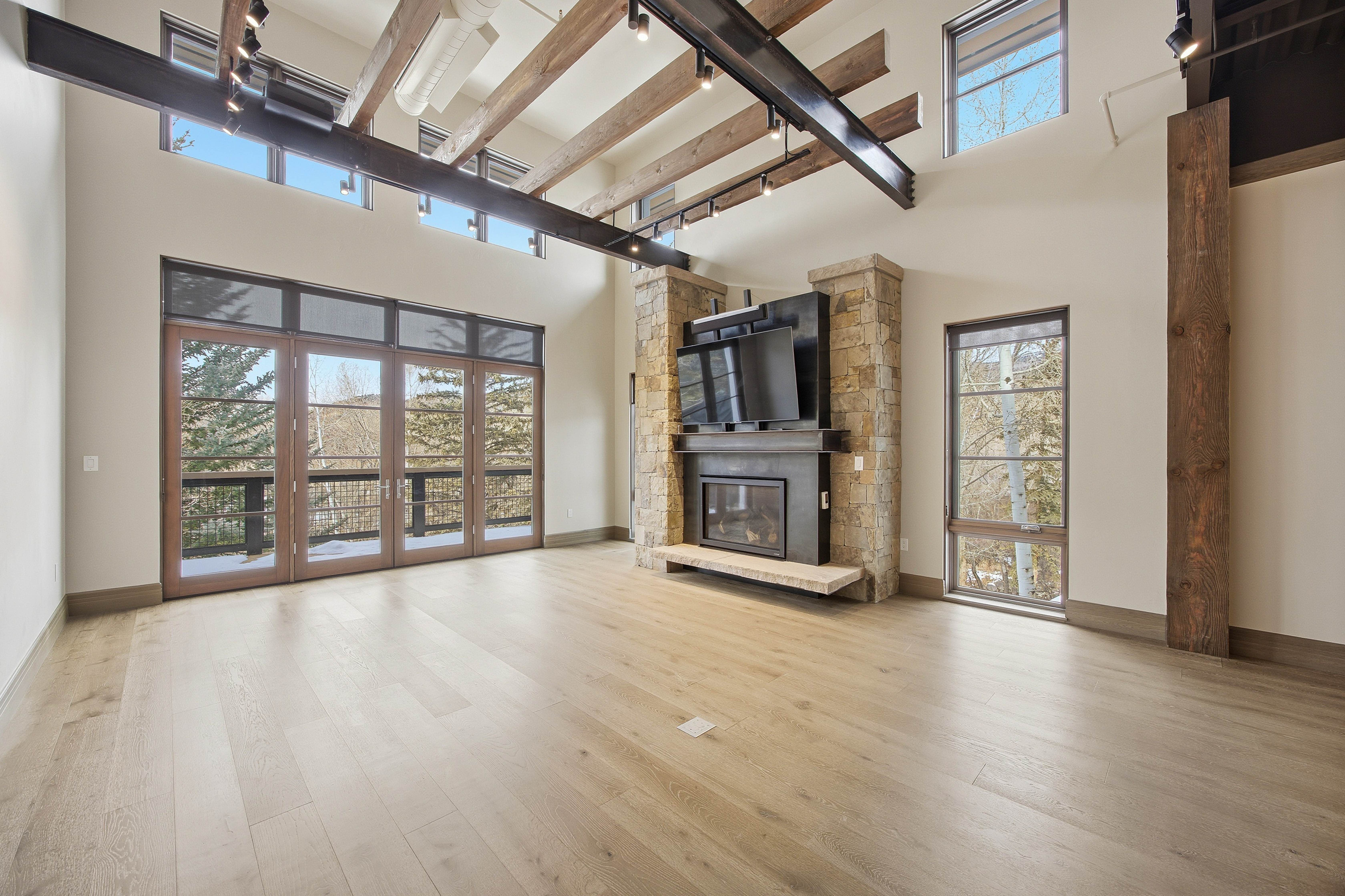 This is a spacious living room featuring a stone fireplace with a mounted television, large windows and doors providing ample natural light, and light wood flooring. The room has high ceilings with exposed wooden beams and metal supports, creating a modern yet rustic aesthetic. The overall impression is one of warmth and sophistication.