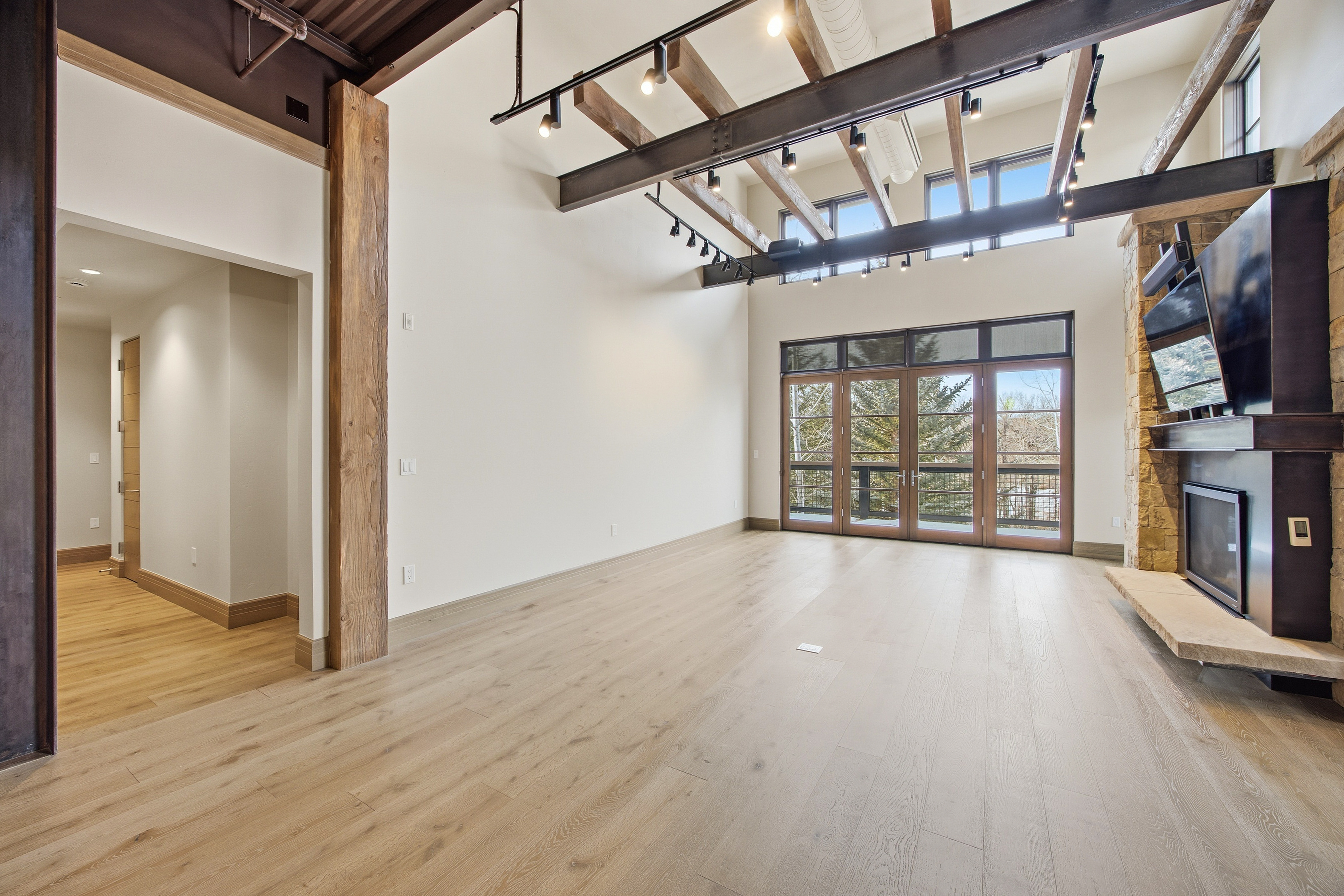 This is a spacious living room featuring light wood flooring and high ceilings with exposed beams and track lighting. A stone fireplace with a dark mantle and mounted television anchors one side of the room, while large glass doors offer views of the outdoors. The room is bright and airy, creating a modern and inviting atmosphere.