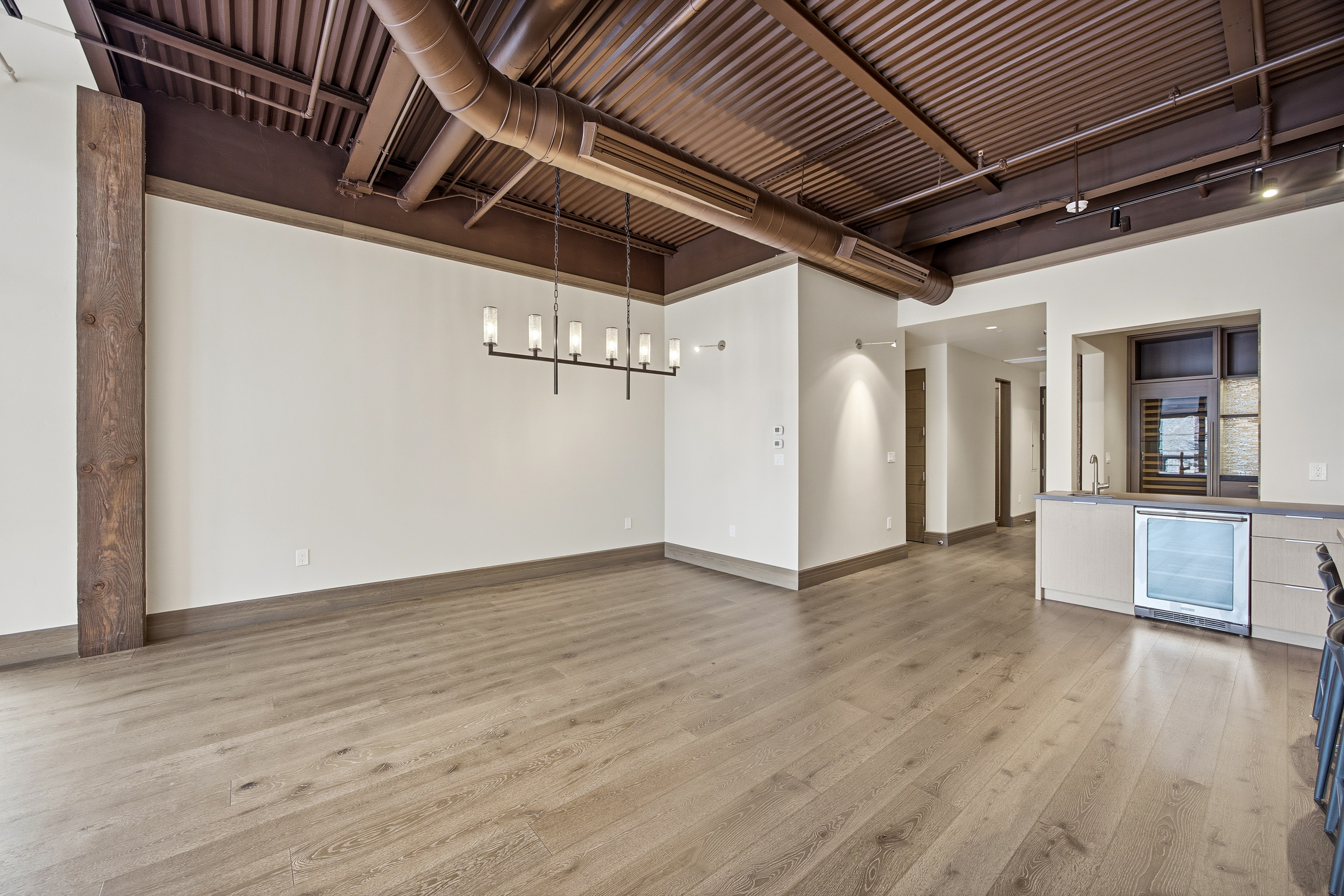 This interior shot showcases a spacious living room with modern industrial design elements. The room features light wood flooring, exposed ductwork and a corrugated metal ceiling painted in a dark bronze color. A contemporary chandelier hangs above the center of the room, and a built-in bar area with a beverage refrigerator is visible in the background.