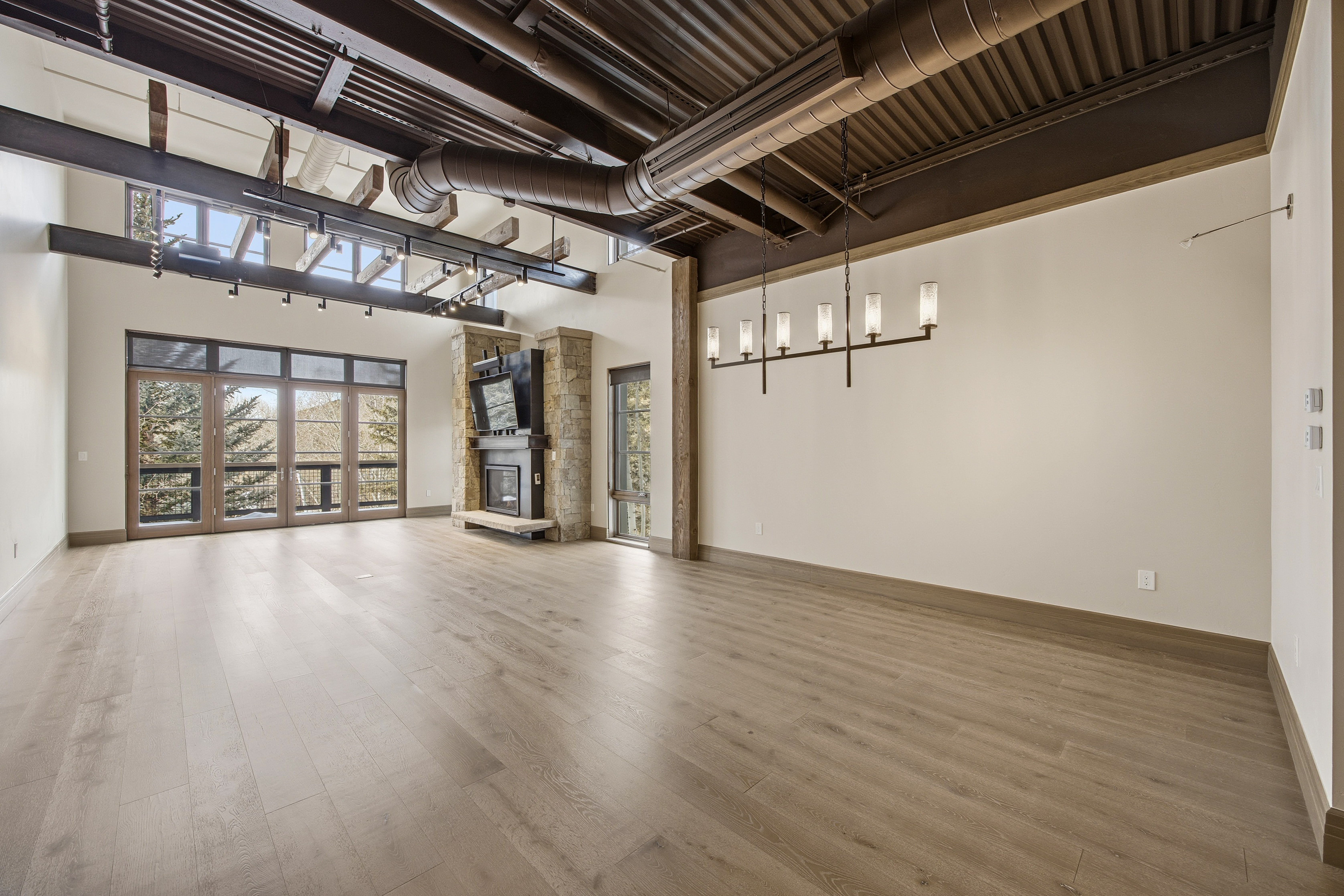 This is a spacious living room featuring light wood flooring and a modern industrial aesthetic. The room has a stone fireplace with a mounted television above it, large windows and doors providing ample natural light, and exposed ductwork and beams on the ceiling. A contemporary chandelier hangs over the room, adding a touch of elegance to the otherwise raw and open space.