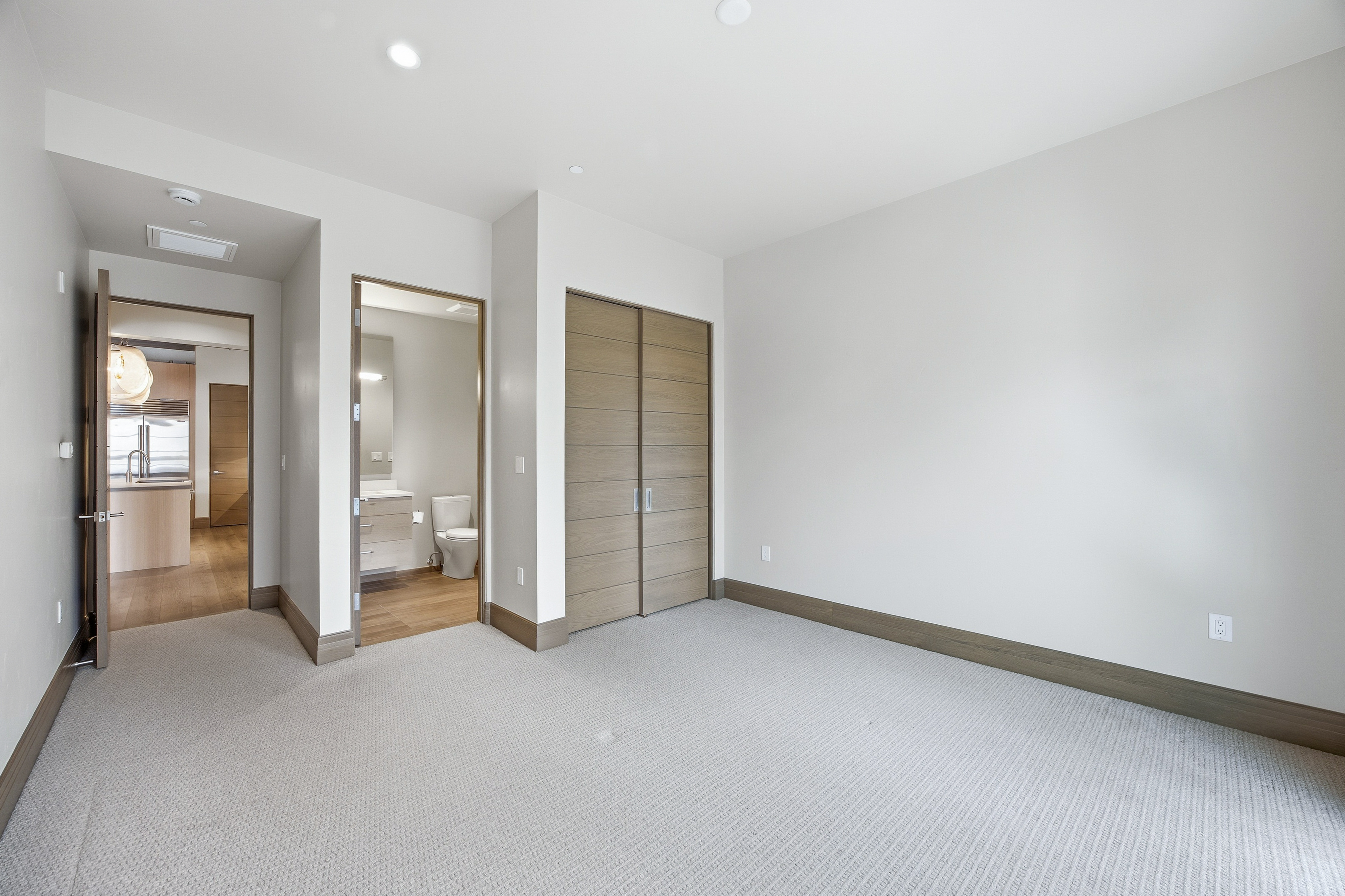 This is a well-lit primary bedroom featuring neutral tones and a minimalist design. The room has light gray carpet, light gray walls, and wood trim. There is a closet with sliding doors, an ensuite bathroom, and an open doorway leading to another room, possibly a kitchen or living area.