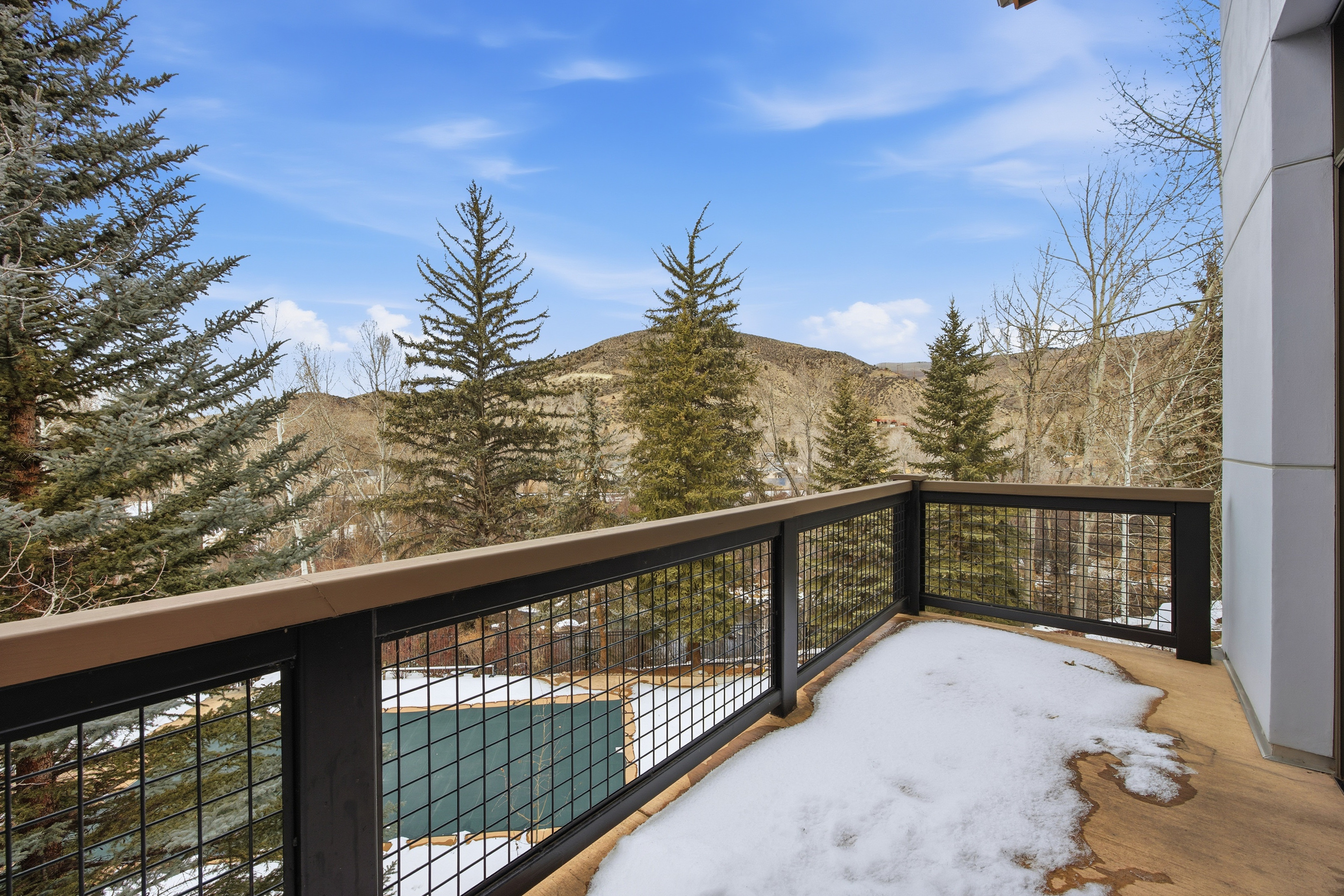 This image showcases a balcony with a view of a landscape featuring trees and a distant mountain under a partly cloudy sky. The balcony has a wooden deck partially covered in snow and a black metal railing with a wooden top. Below, a pool is visible, suggesting a luxurious outdoor living space.