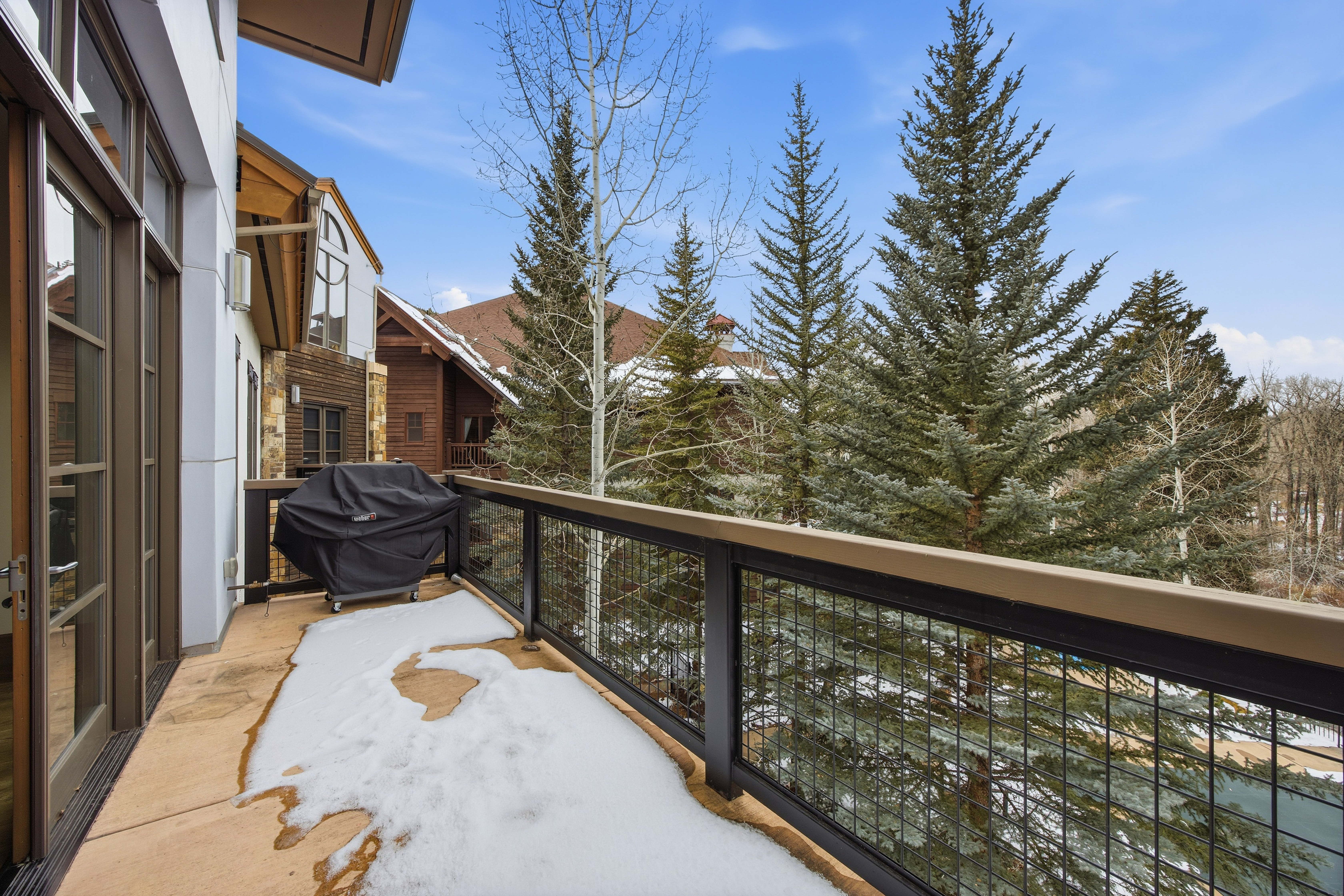 This image showcases a balcony with a metal railing and a wooden top rail, partially covered in snow. A covered grill sits on the balcony, and beyond the railing are evergreen trees and a glimpse of other houses. The view suggests a mountain or wooded setting, adding to the property's appeal.
