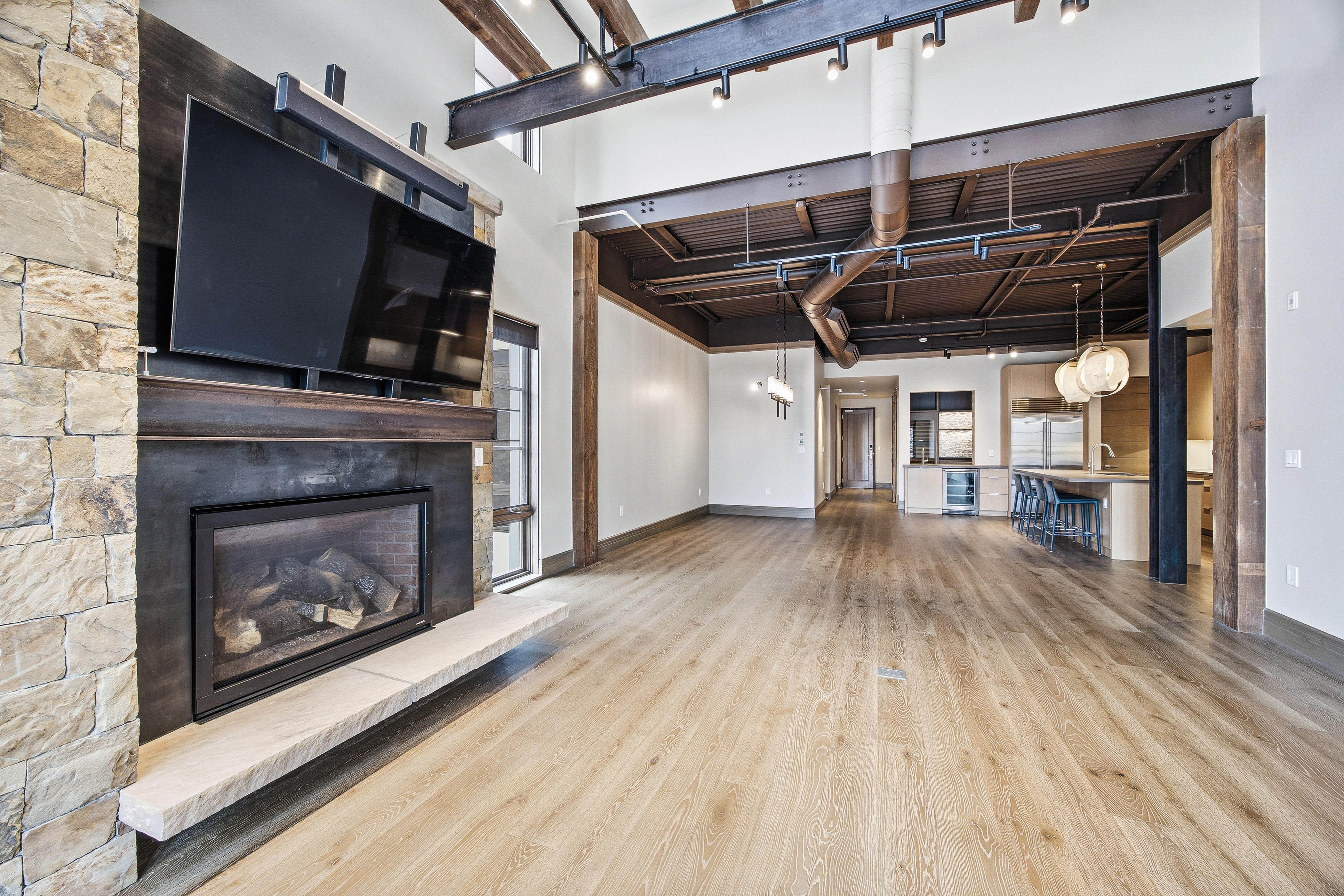 This is a spacious living room featuring a stone fireplace with a large television mounted above. The room has an open floor plan that flows into the kitchen area, with exposed beams and ductwork adding an industrial-chic touch. Light wood flooring and ample natural light create a bright and inviting atmosphere.