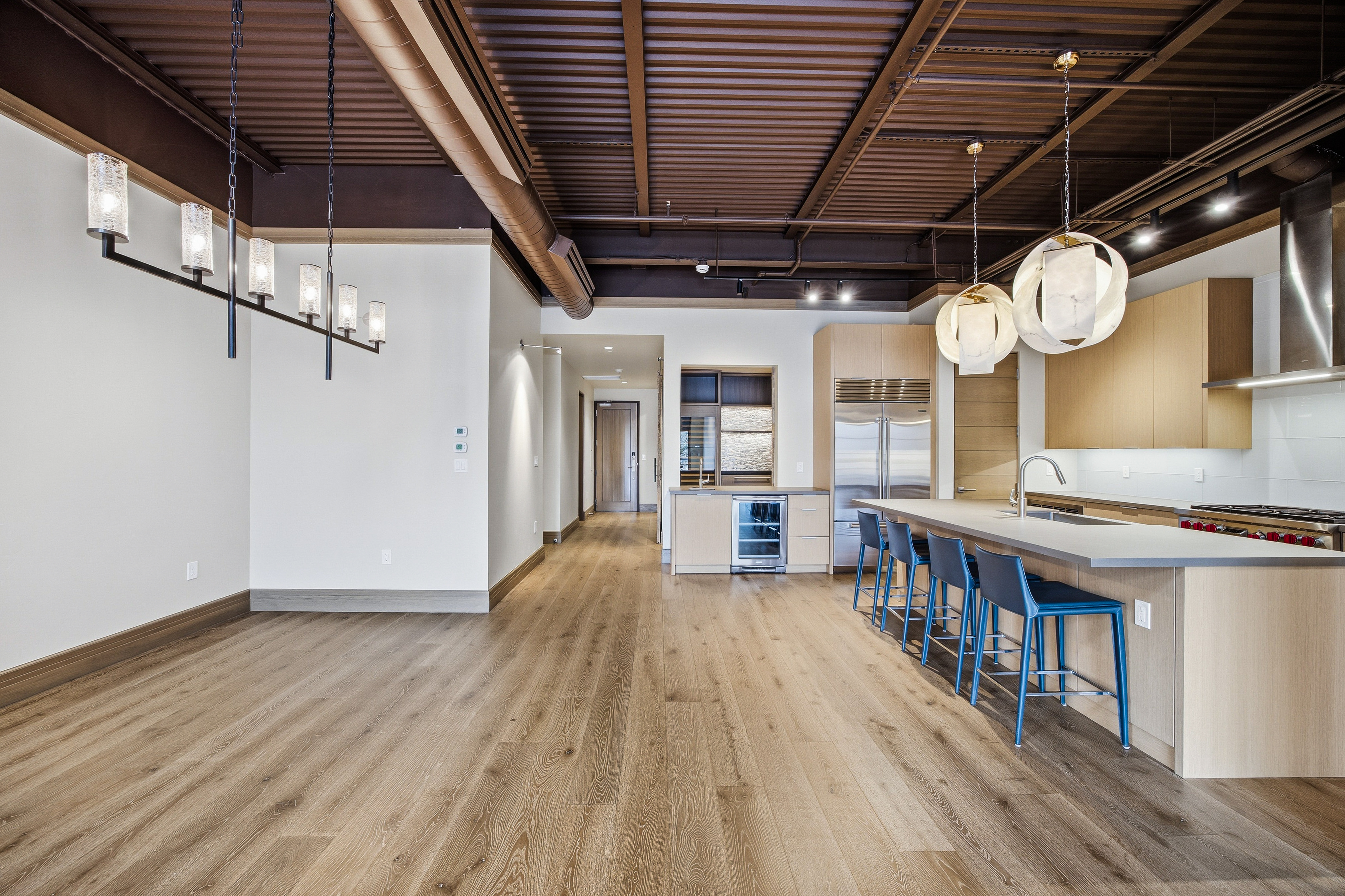 This interior shot showcases a modern kitchen and living area with an open floor plan. The kitchen features a large island with seating, stainless steel appliances, and light wood cabinetry. The living area is spacious with hardwood floors and contemporary lighting fixtures, creating a bright and inviting atmosphere.