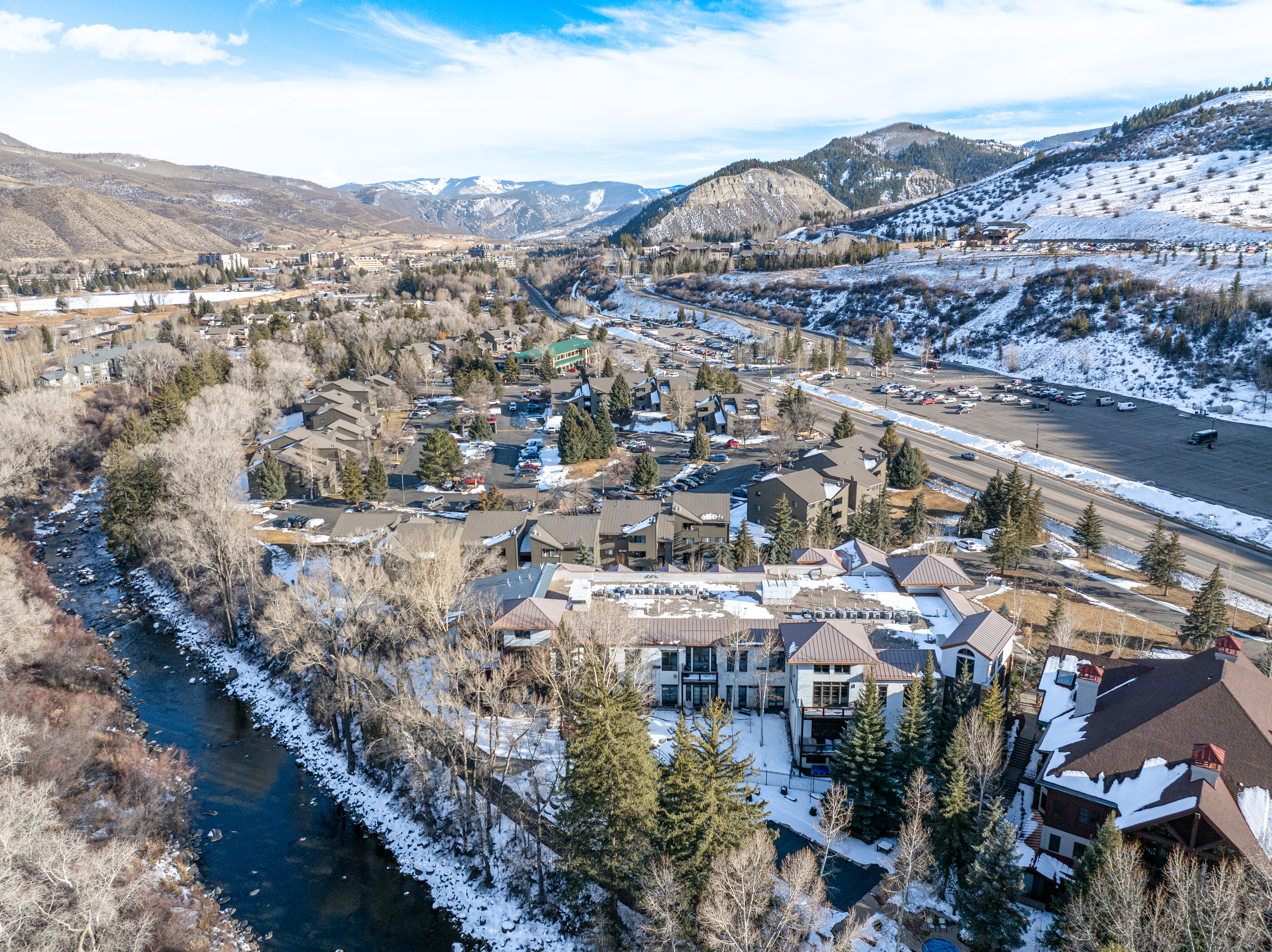 This aerial view showcases a complex of residential buildings nestled alongside a river, with snow-dusted mountains in the background. The buildings feature a mix of architectural styles, with some having brown roofs and others with lighter, possibly metal, roofing. A road and parking area are visible, indicating convenient access, and the surrounding landscape includes a mix of trees and snow-covered ground, creating a serene and picturesque setting.