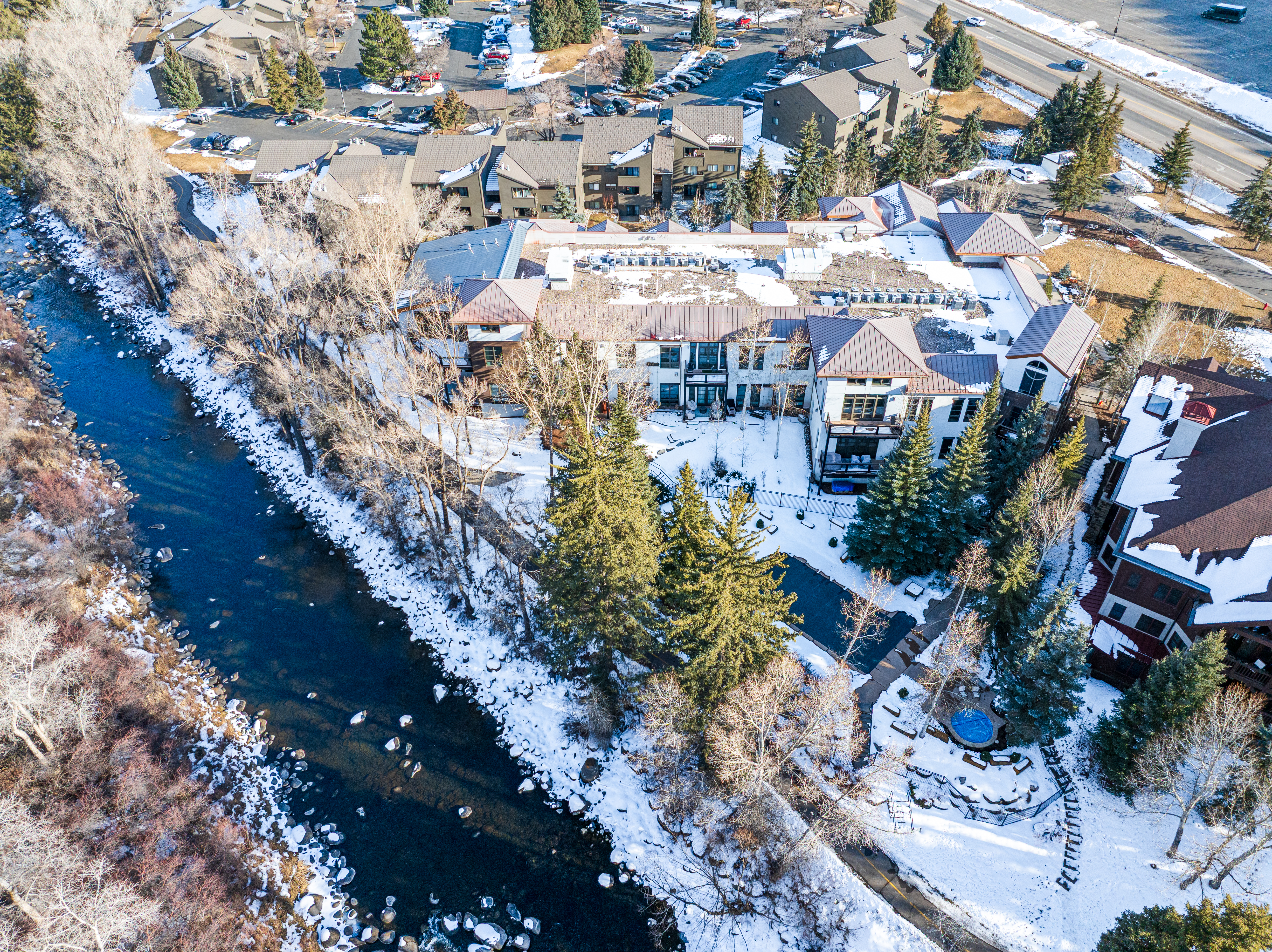This aerial view showcases a multi-unit residential building complex situated alongside a river, partially covered in snow. The buildings feature a mix of flat and pitched roofs, with a modern architectural style. The surrounding landscape includes mature trees, a pathway, and a glimpse of a nearby road, creating a serene yet accessible environment.