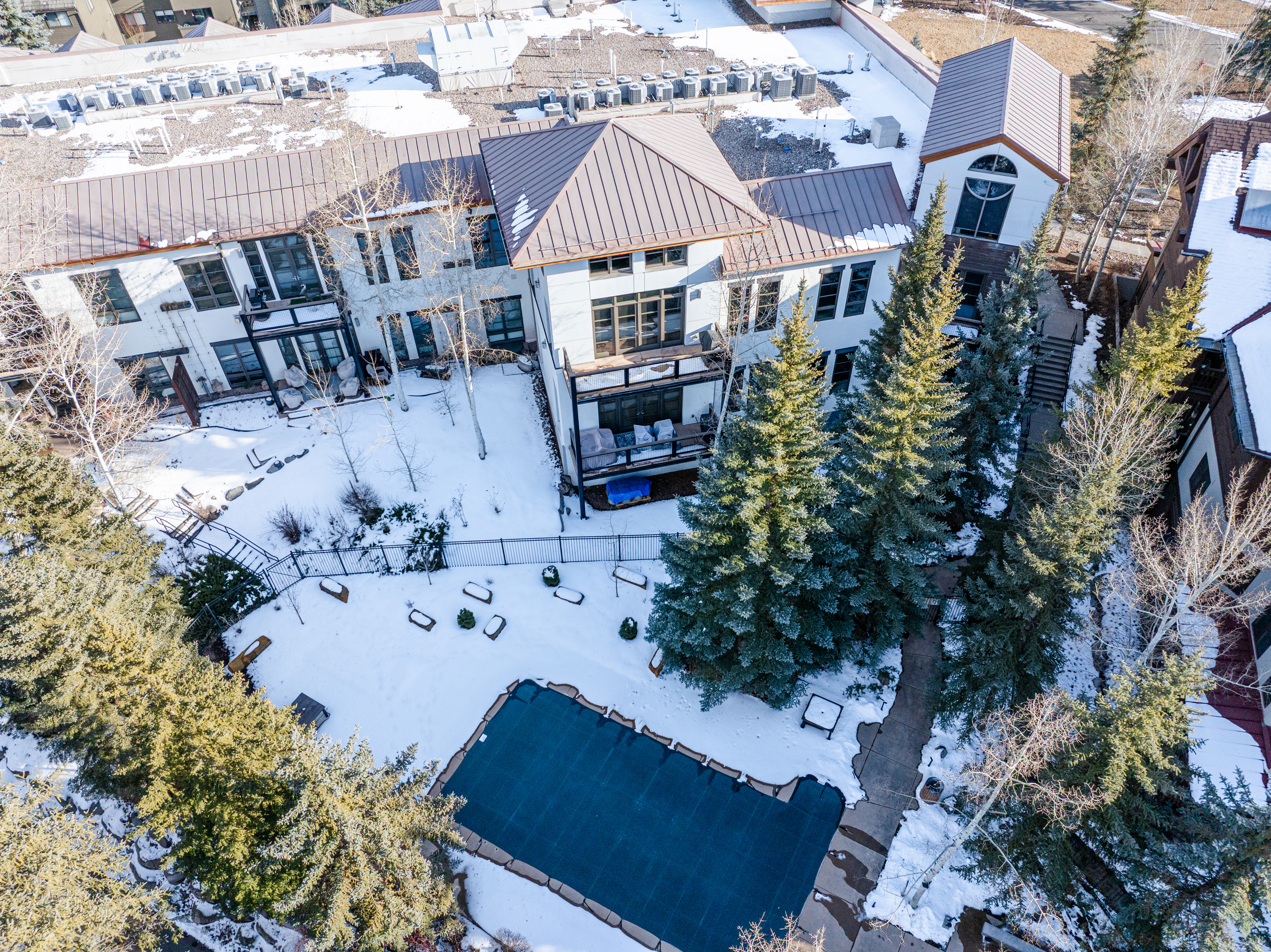 This aerial shot showcases a luxurious property with a metal roof, white exterior, and multiple windows. The backyard features a covered pool, mature evergreen trees, and a patio area, all dusted with snow. The perspective gives a comprehensive view of the property's layout and outdoor amenities.