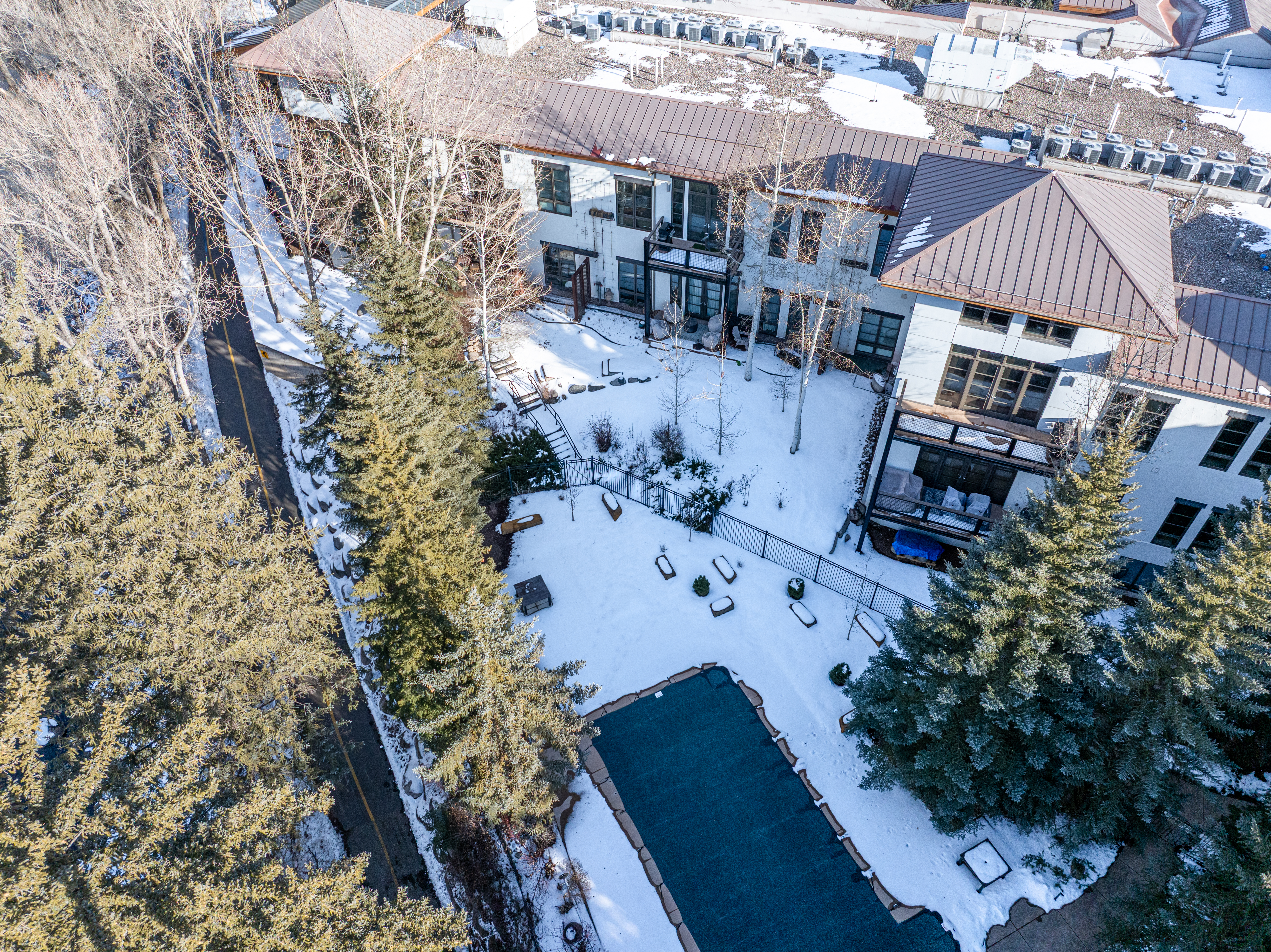 This aerial shot showcases a luxurious multi-story home with a distinctive brown roof and light-colored exterior walls. The property features a covered pool, a well-maintained yard with mature trees, and a pathway alongside the building. The surrounding area is partially covered in snow, adding a serene touch to the landscape.
