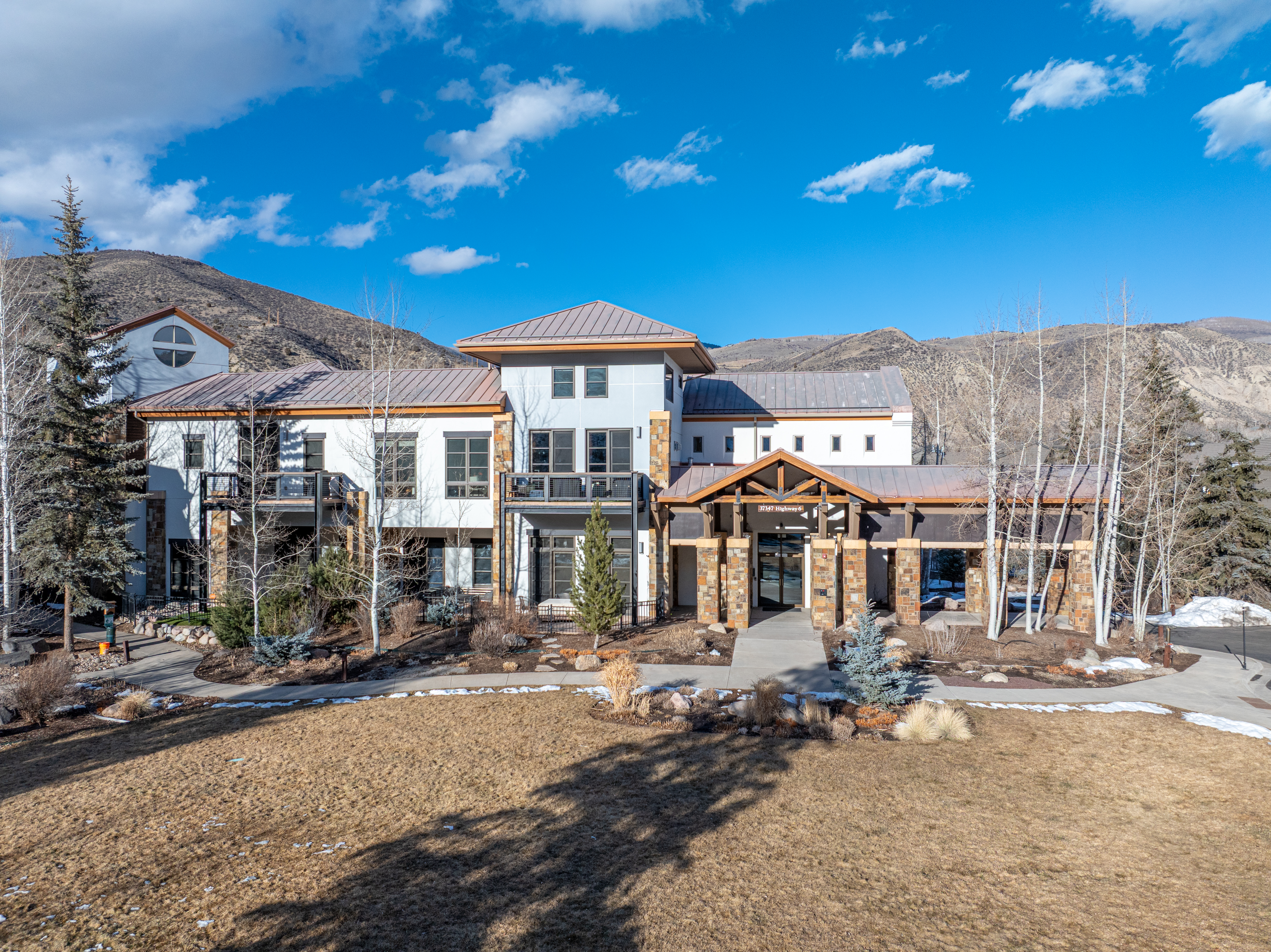 This is a front exterior view of a multi-story building with a modern architectural style. The building features a combination of white and gray exterior walls, accented by stone pillars and a copper-colored roof. The landscape includes a well-maintained lawn, trees, and decorative shrubbery, creating an inviting and upscale appearance.