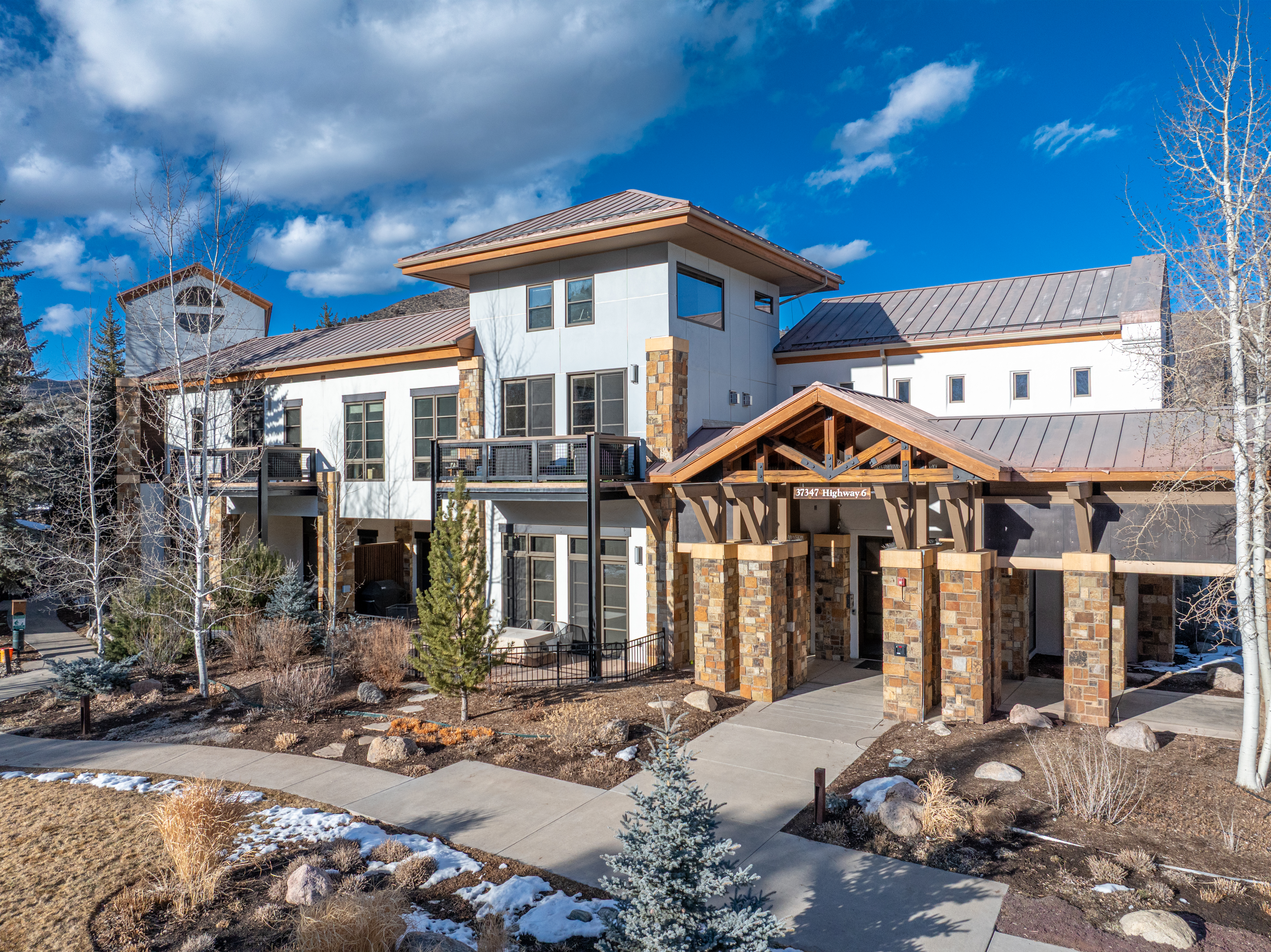This is a welcoming entryway to a modern building, featuring a covered entrance with wooden beams and stone pillars. The building has a contemporary design with a mix of light-colored siding and stone accents. A well-maintained walkway leads to the entrance, surrounded by landscaping with small trees and shrubs, creating an inviting first impression.