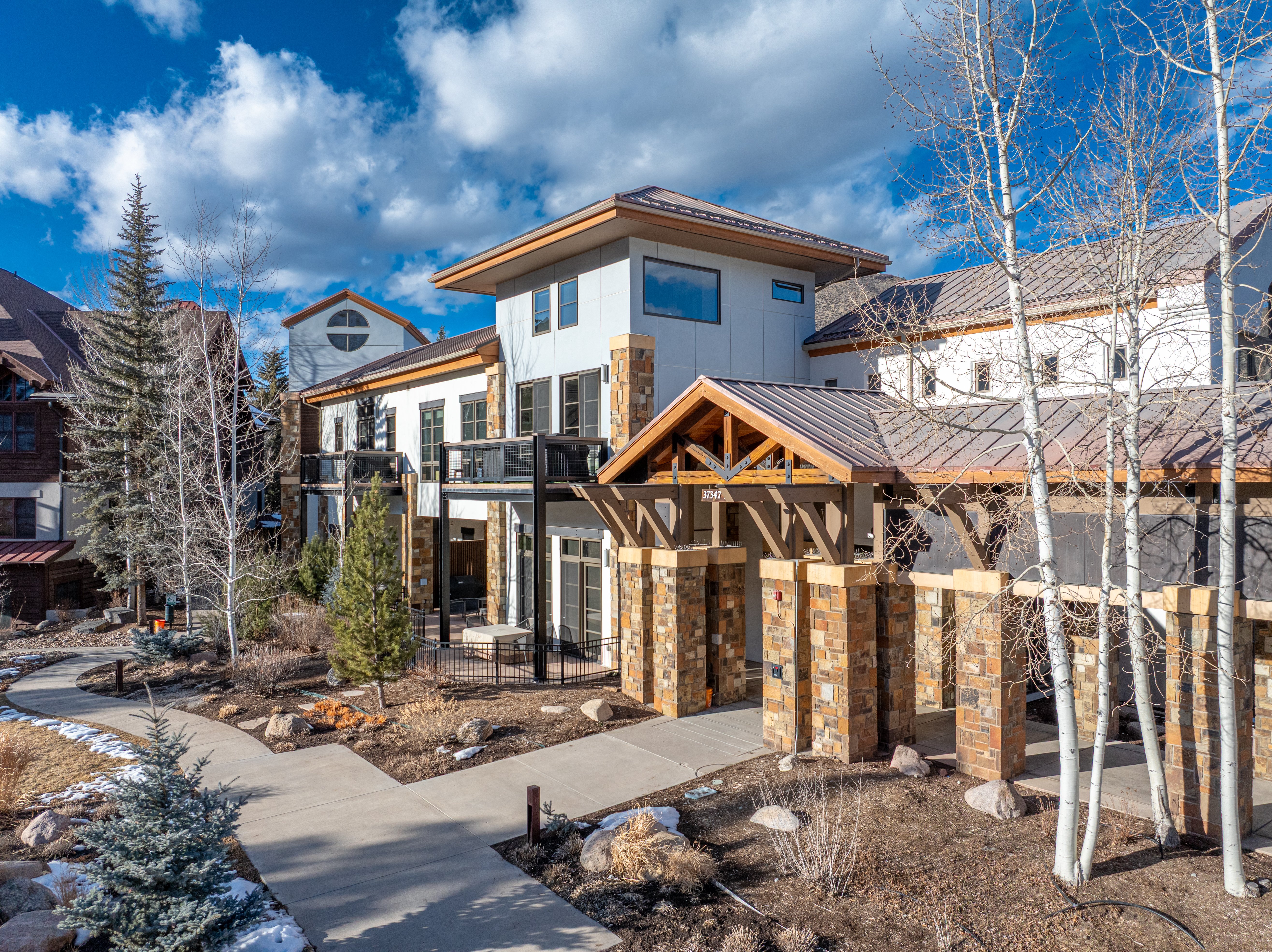 This is a front exterior view of a modern multi-story building with a unique architectural design. The building features a combination of light-colored siding, stone accents, and a distinctive covered entryway with wooden beams and stone pillars. A paved walkway leads to the entrance, surrounded by landscaping with trees and shrubs, creating an inviting and upscale impression.