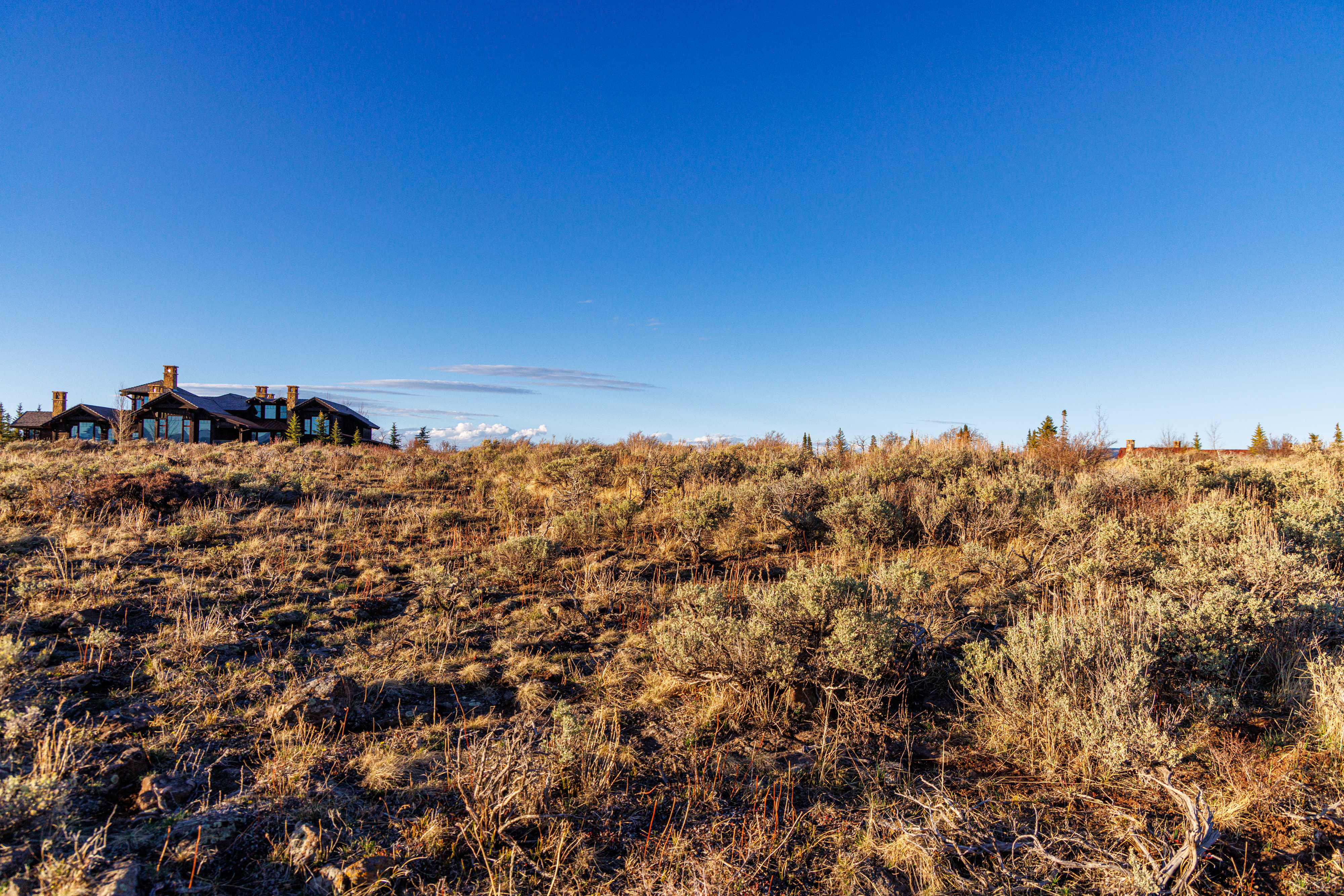 This wide-angle shot captures a rustic, multi-gabled luxury home nestled on a vast, arid landscape under a clear, deep blue sky. The foreground is dominated by native sagebrush and dry grasses, emphasizing the expansive, secluded nature of the property. The architectural style features natural stone and dark wood siding, blending harmoniously with the rugged, high-desert environment.