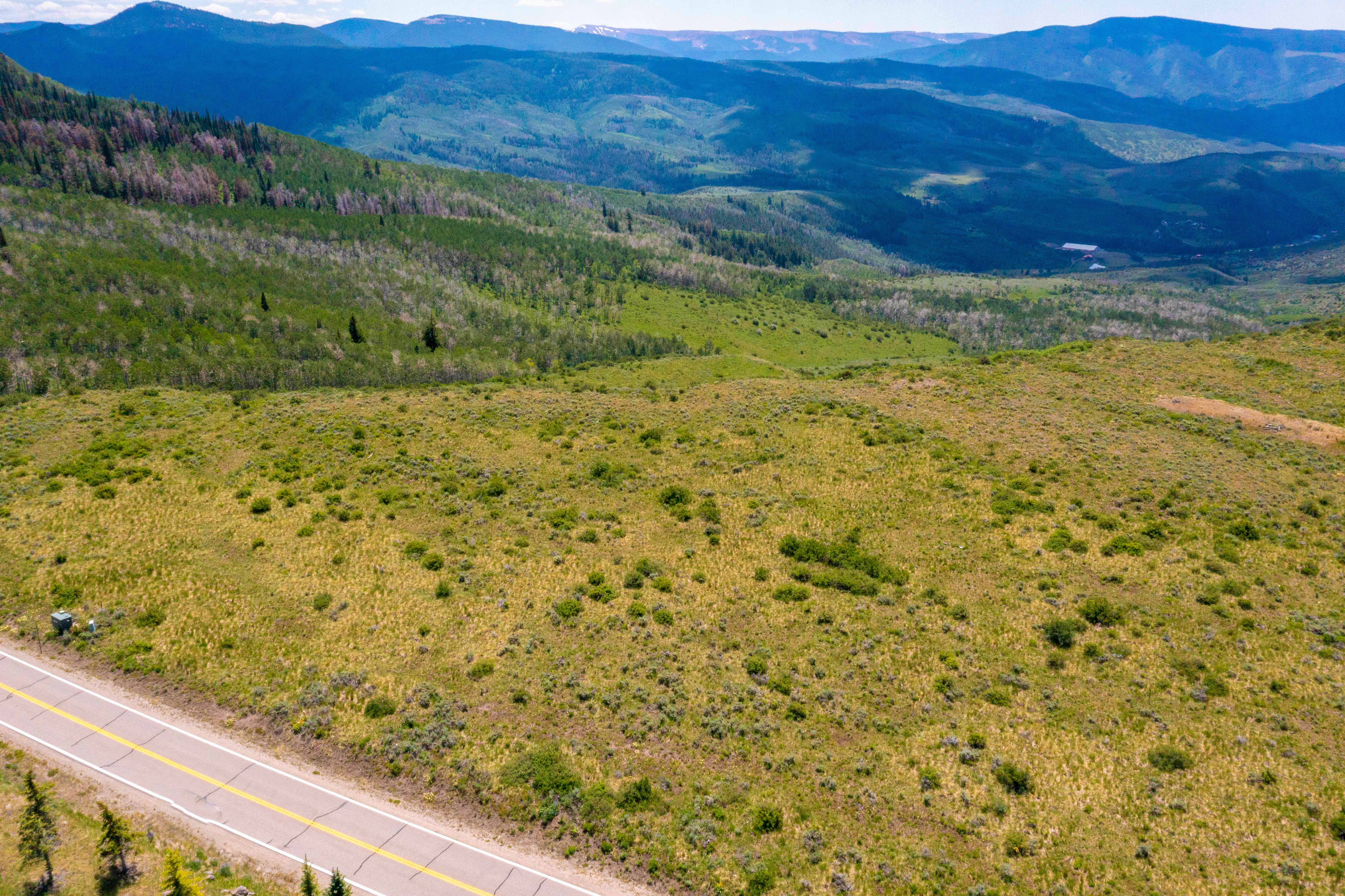 This high-angle aerial shot captures a vast, open landscape featuring a paved road winding through rolling, grassy terrain dotted with low-lying shrubs. In the background, a dense forest transitions into majestic, layered mountain ranges under a clear blue sky. The perspective emphasizes the expansive, natural beauty and the secluded, scenic location of the property.
