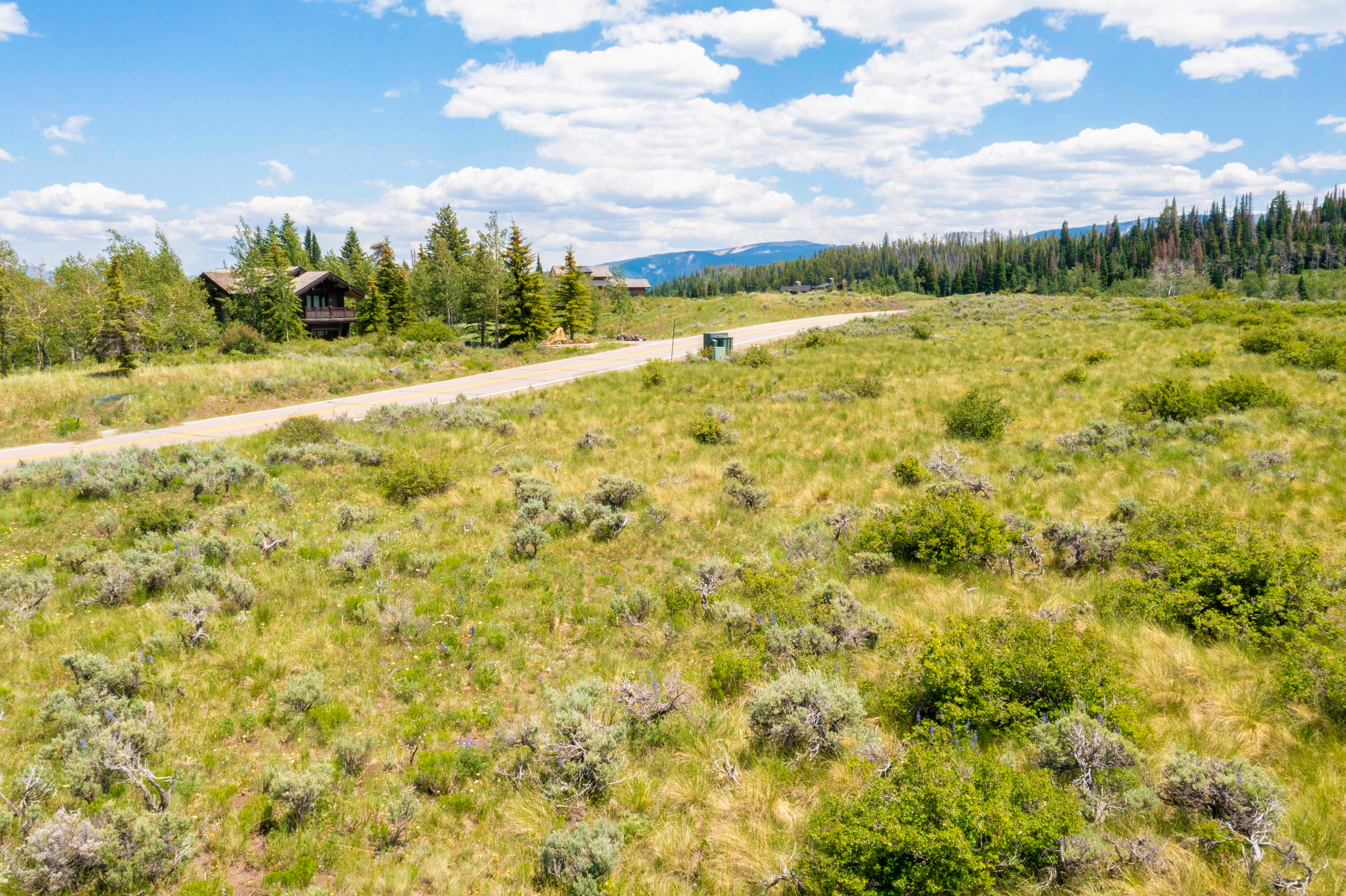 This aerial perspective captures a vast, open landscape characterized by rolling fields of sagebrush and native grasses, set against a backdrop of distant mountains and a clear, blue sky with scattered clouds. A paved road cuts through the foreground, leading toward a secluded wooden cabin nestled among evergreen trees. The scene conveys a sense of expansive, peaceful wilderness, ideal for highlighting the natural beauty and privacy of the property's location.