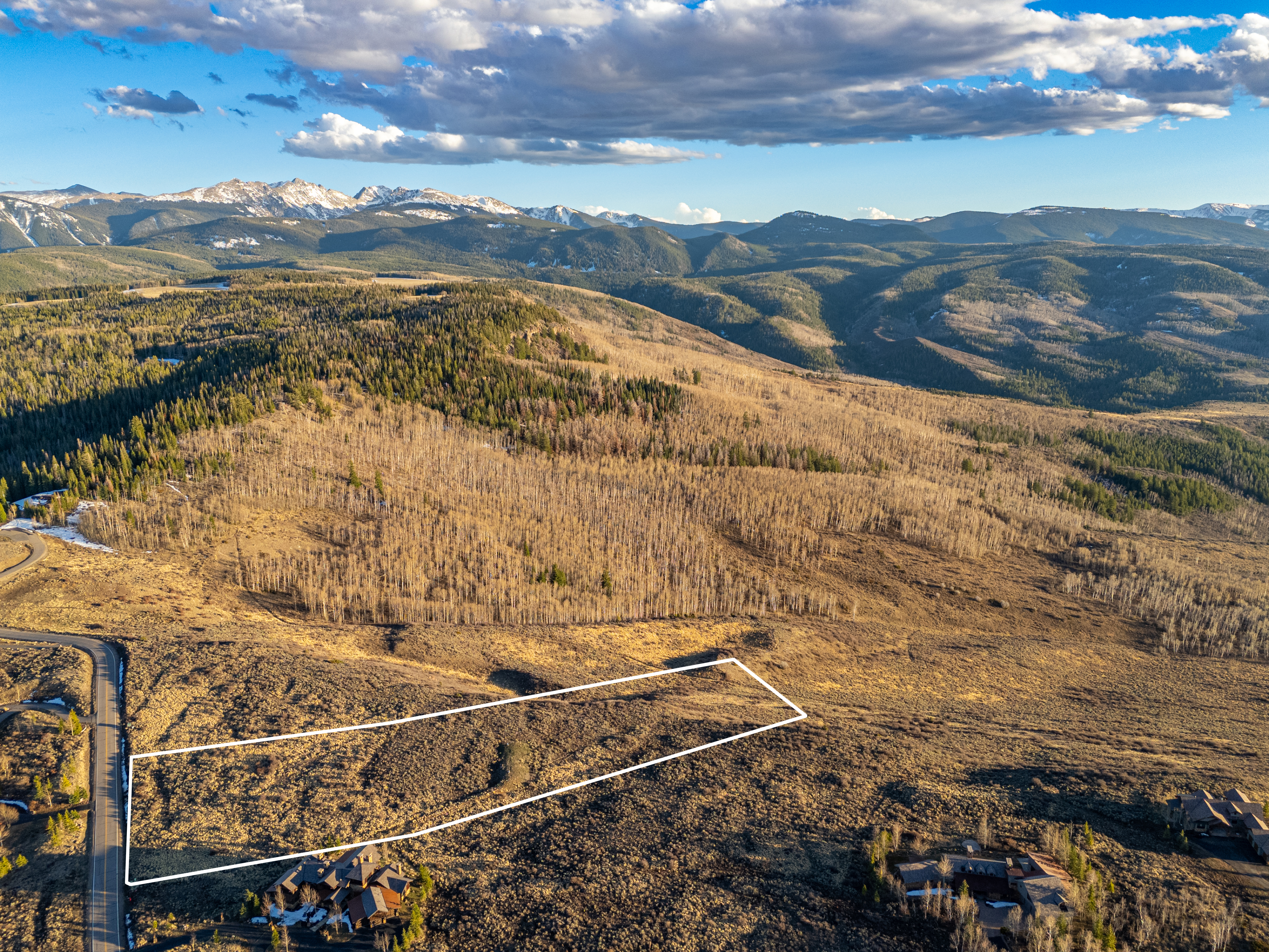 This high-angle aerial shot captures a vast, undeveloped plot of land outlined in white, set against a breathtaking backdrop of rugged, snow-capped mountains and dense forest. The landscape features rolling, golden-hued terrain typical of high-altitude regions, with a winding road and nearby luxury residential properties providing context for the location. The perspective emphasizes the expansive scale and serene, secluded nature of the property, making it an ideal candidate for a custom mountain retreat.