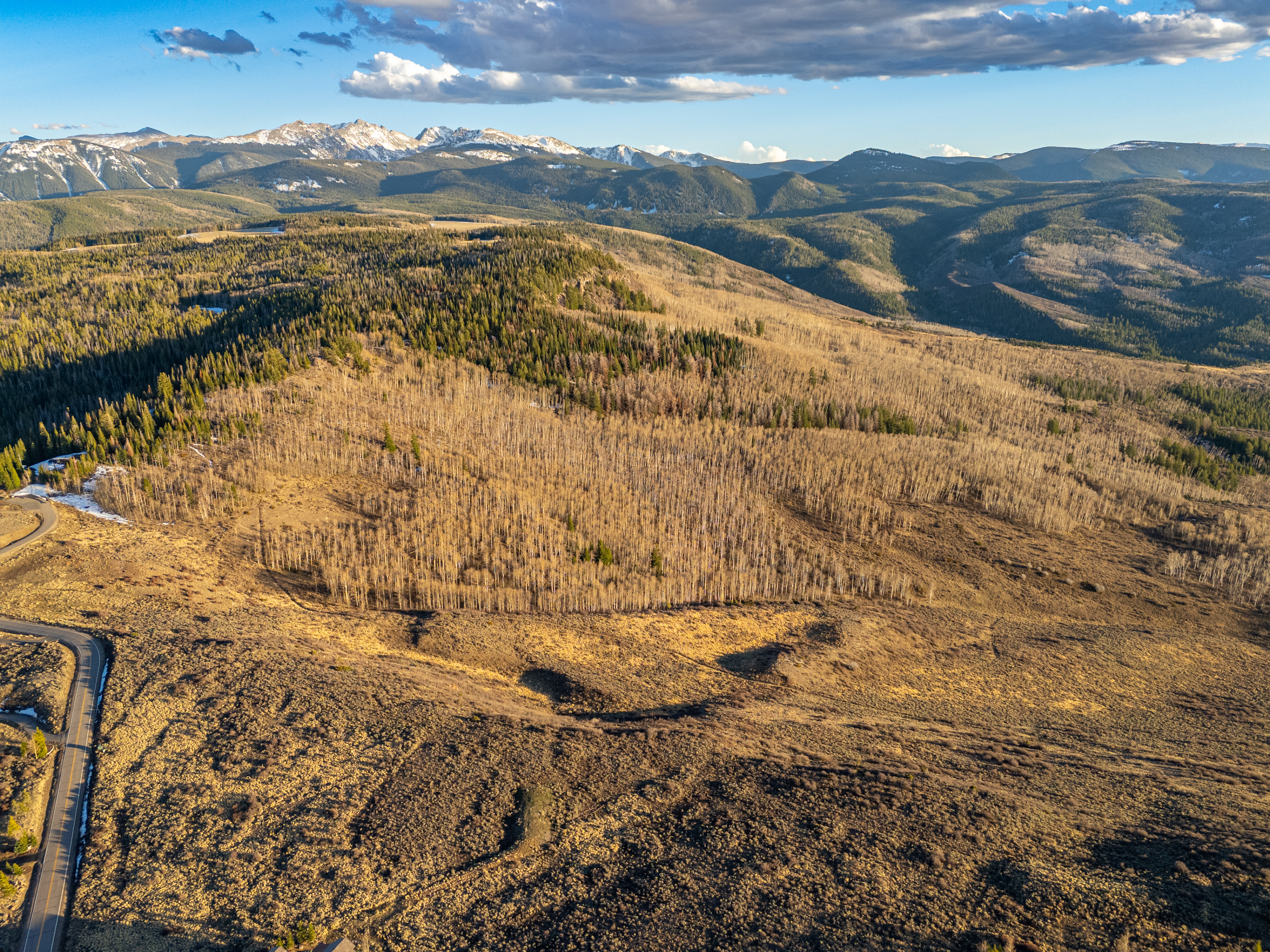 This stunning aerial view captures a vast, rugged landscape featuring a dense forest of aspen trees transitioning into evergreen pines against a backdrop of majestic, snow-capped mountains. A winding paved road snakes through the foreground, emphasizing the property's remote and scenic location. The golden hues of the late afternoon sun cast long shadows, highlighting the dramatic topography and natural beauty of the terrain.