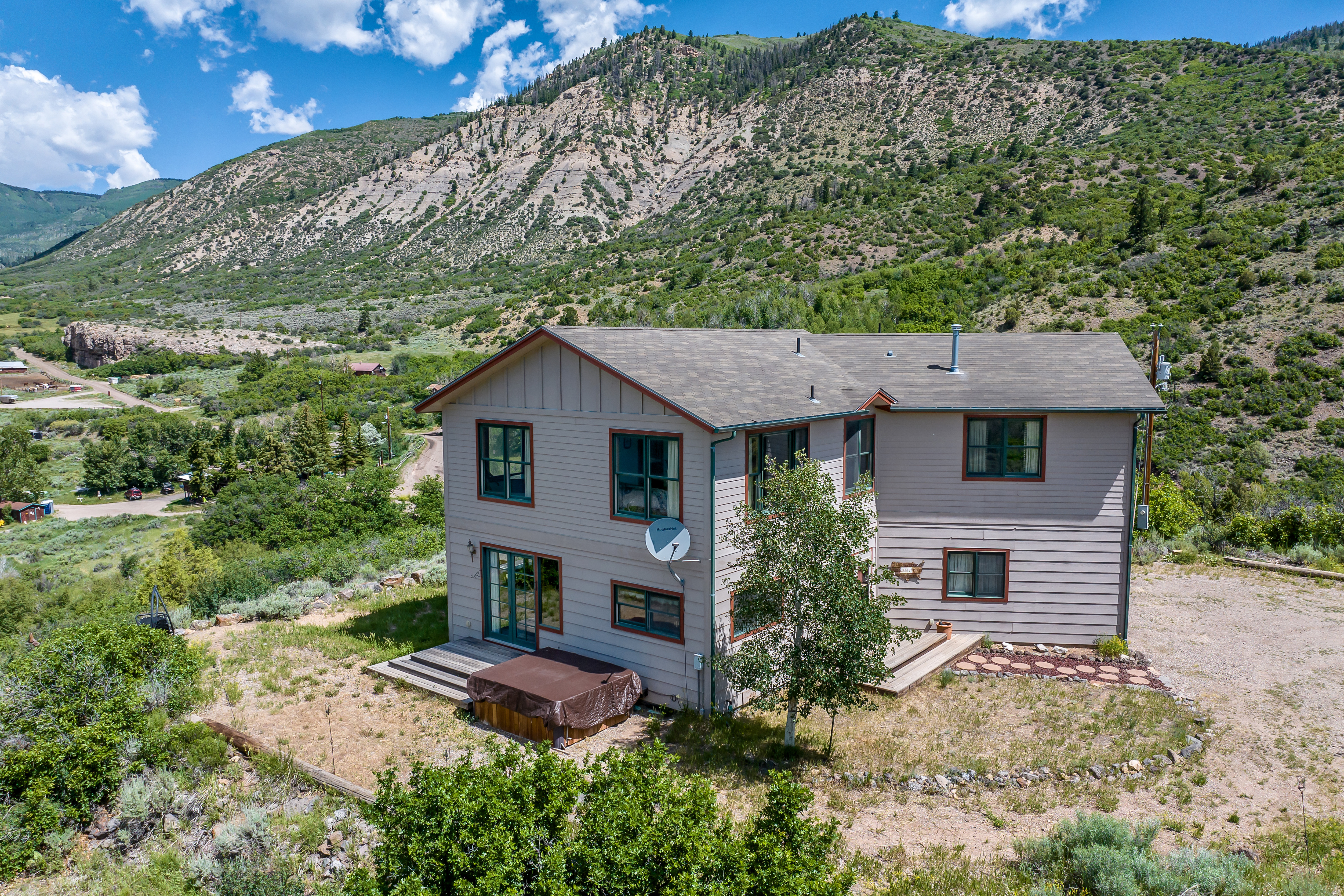 This aerial perspective captures a two-story, light-colored wood-sided home nestled against a dramatic, rugged mountain backdrop. The property features a wooden deck with a hot tub, a natural landscape with native shrubs, and a gravel driveway, offering a serene and secluded mountain retreat atmosphere. The wide-angle shot emphasizes the home's integration into the expansive, scenic environment.