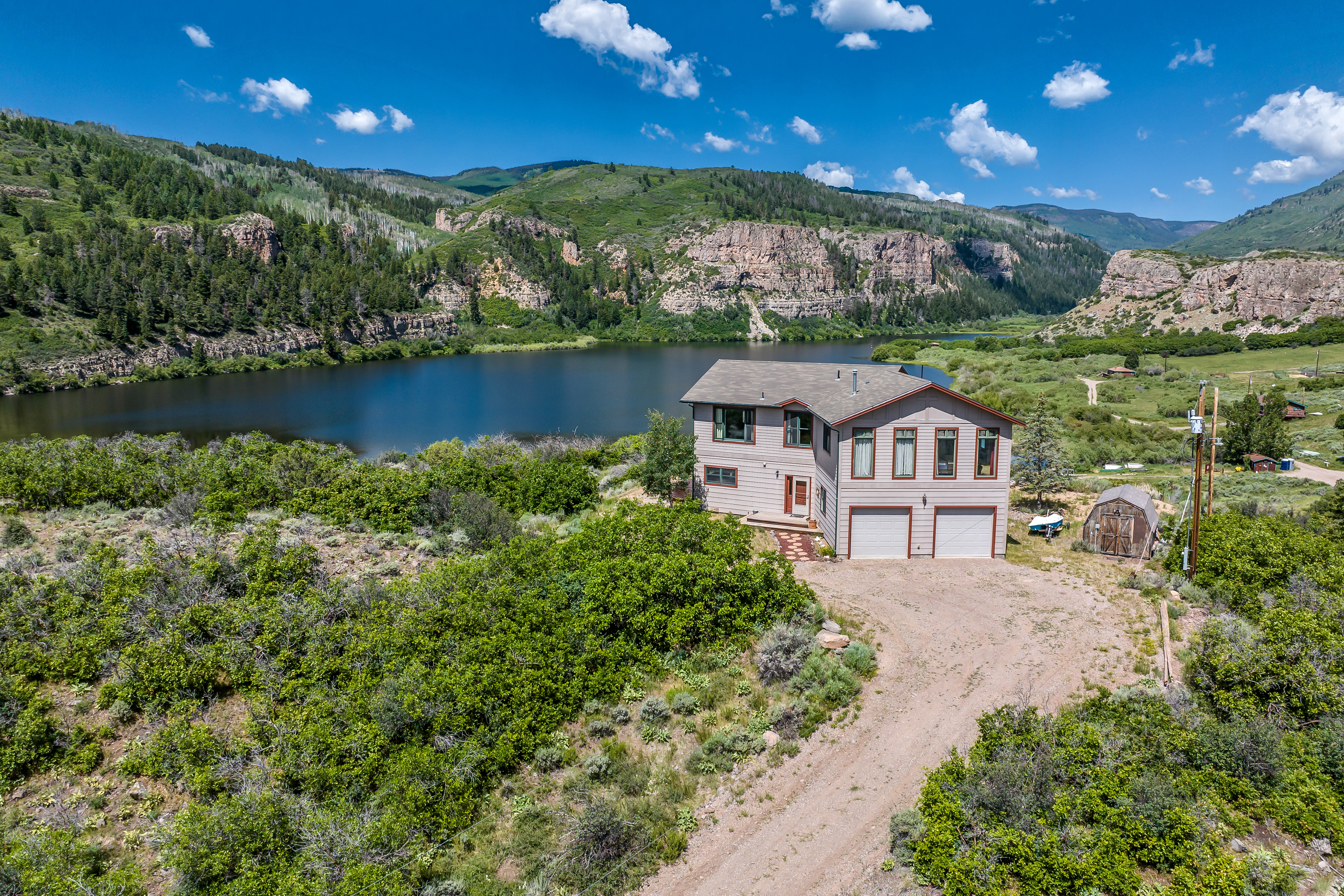 This high-angle aerial shot captures a two-story residential home nestled in a stunning, rugged landscape overlooking a serene lake. The property features a multi-car garage, a gravel driveway, and a small shed, all surrounded by lush green vegetation and dramatic rocky cliffs in the background. The perspective emphasizes the home's secluded, picturesque location, highlighting its integration with the natural environment.