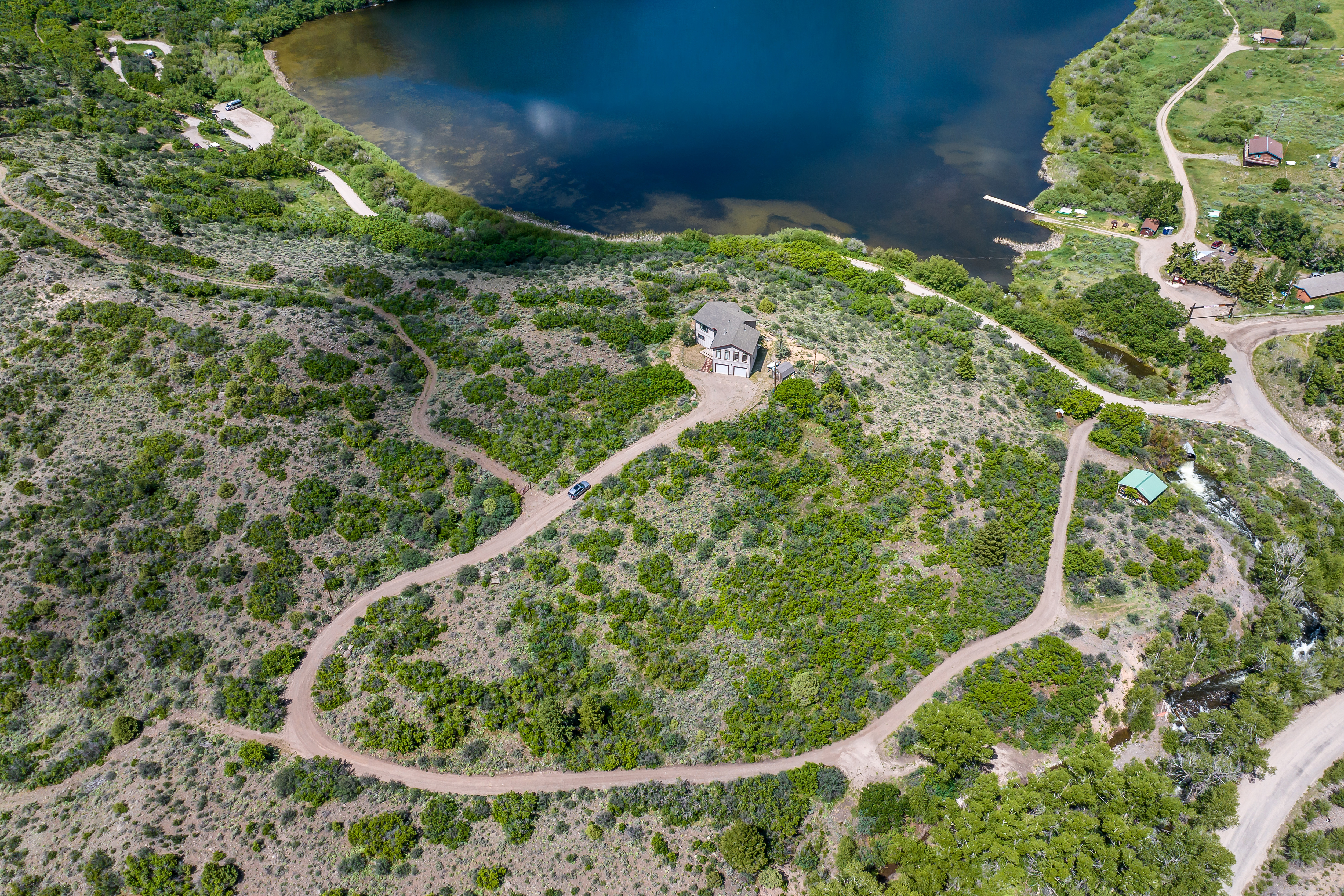 This high-angle aerial view captures a secluded residential property nestled on a hillside overlooking a serene, deep blue lake. The landscape is characterized by rolling, shrub-covered terrain, a winding dirt driveway leading to the home, and a nearby stream flowing through the lower valley. The perspective emphasizes the property's expansive natural setting and its peaceful, remote location.