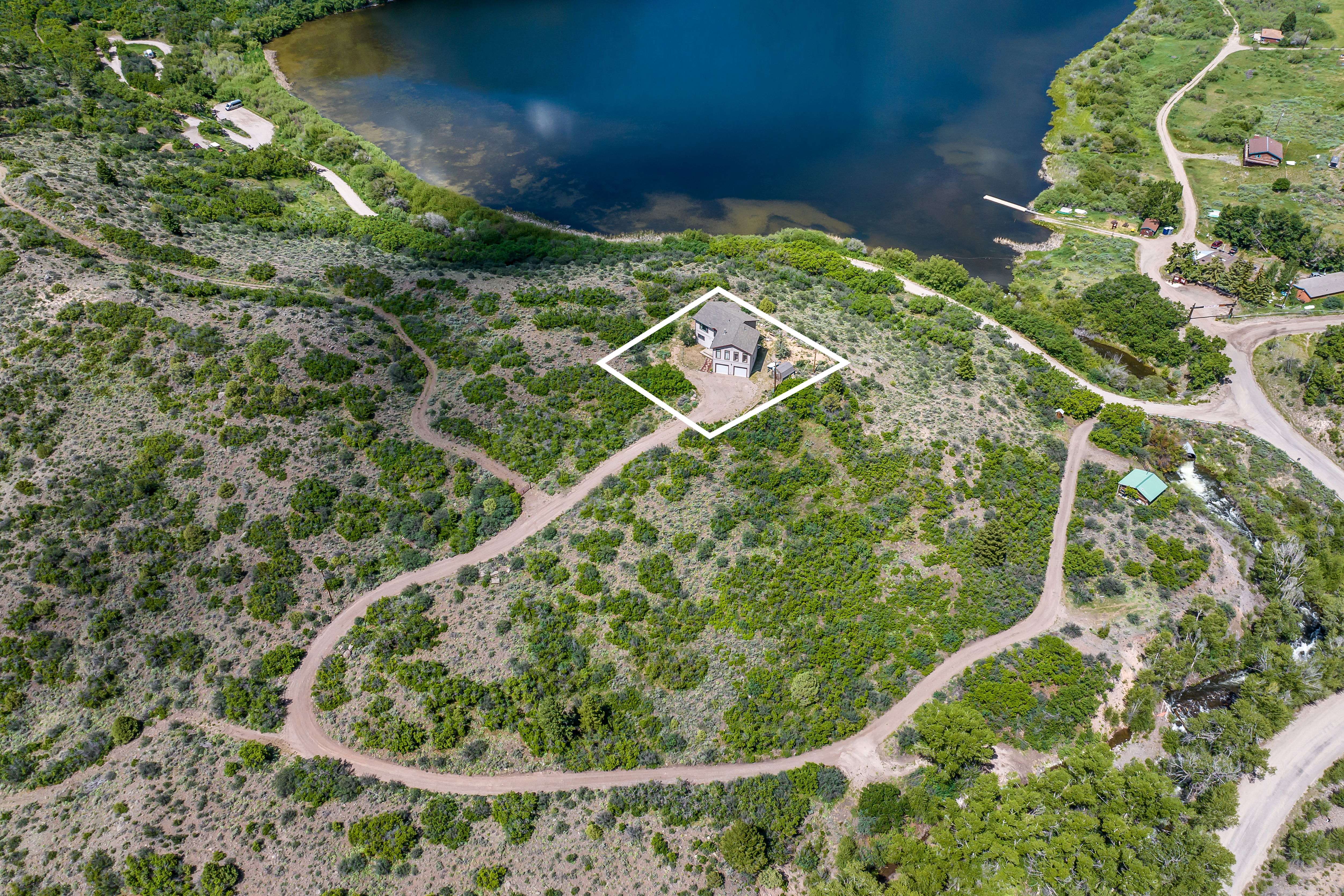 This high-angle aerial shot captures a secluded residential property nestled on a hillside overlooking a large, serene lake. The home is highlighted by a white boundary overlay, showcasing its private driveway and natural surroundings, including dense vegetation and nearby water features. The perspective emphasizes the property's expansive lot and its tranquil, scenic location.