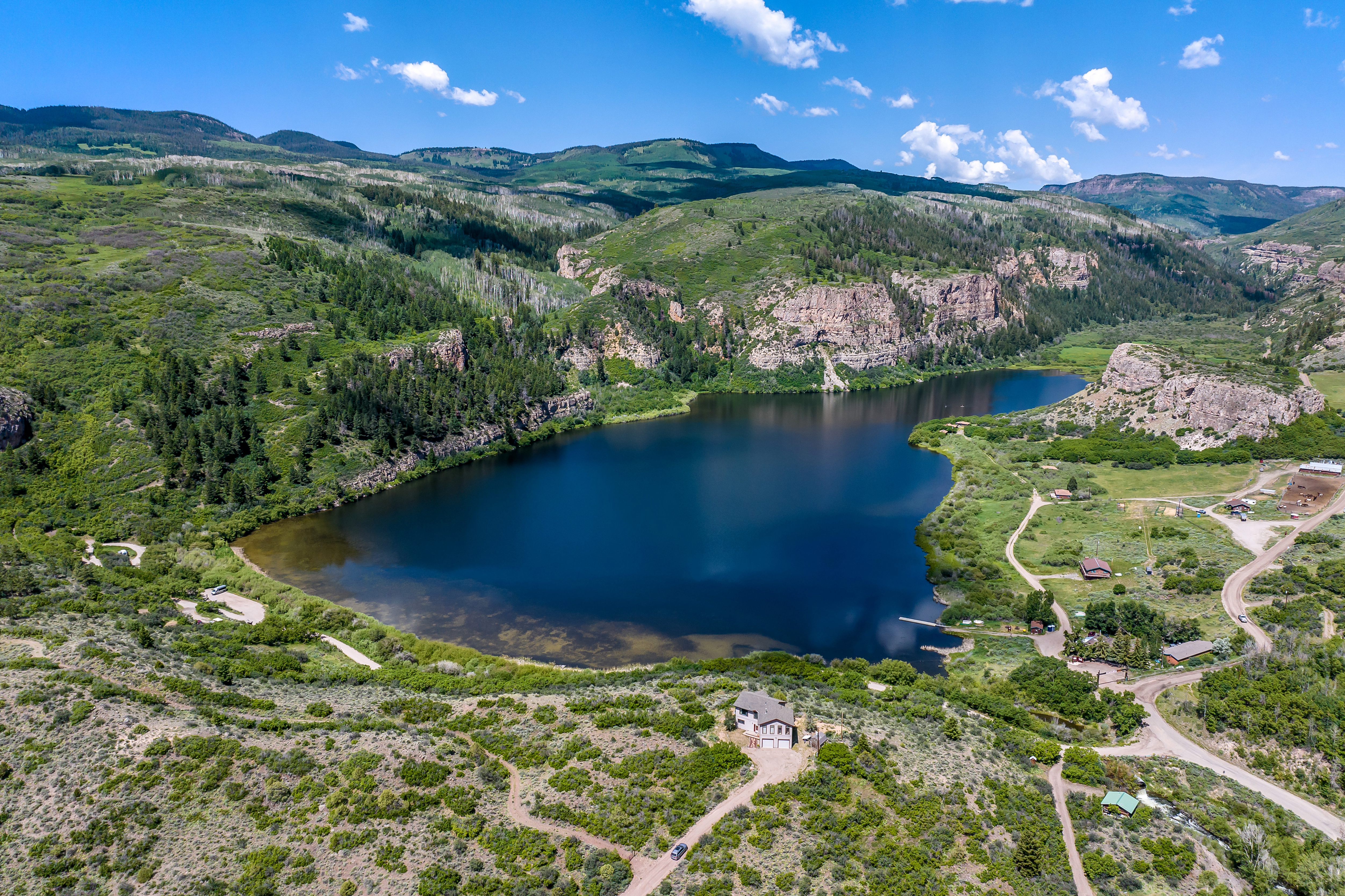 This stunning aerial view captures a secluded mountain property nestled beside a large, deep blue lake surrounded by rugged cliffs and lush, rolling hills. The perspective highlights the expansive natural landscape, showcasing the property's prime waterfront location and proximity to wilderness trails. The scene conveys a sense of peaceful isolation and grand scale, perfect for those seeking a private retreat in nature.