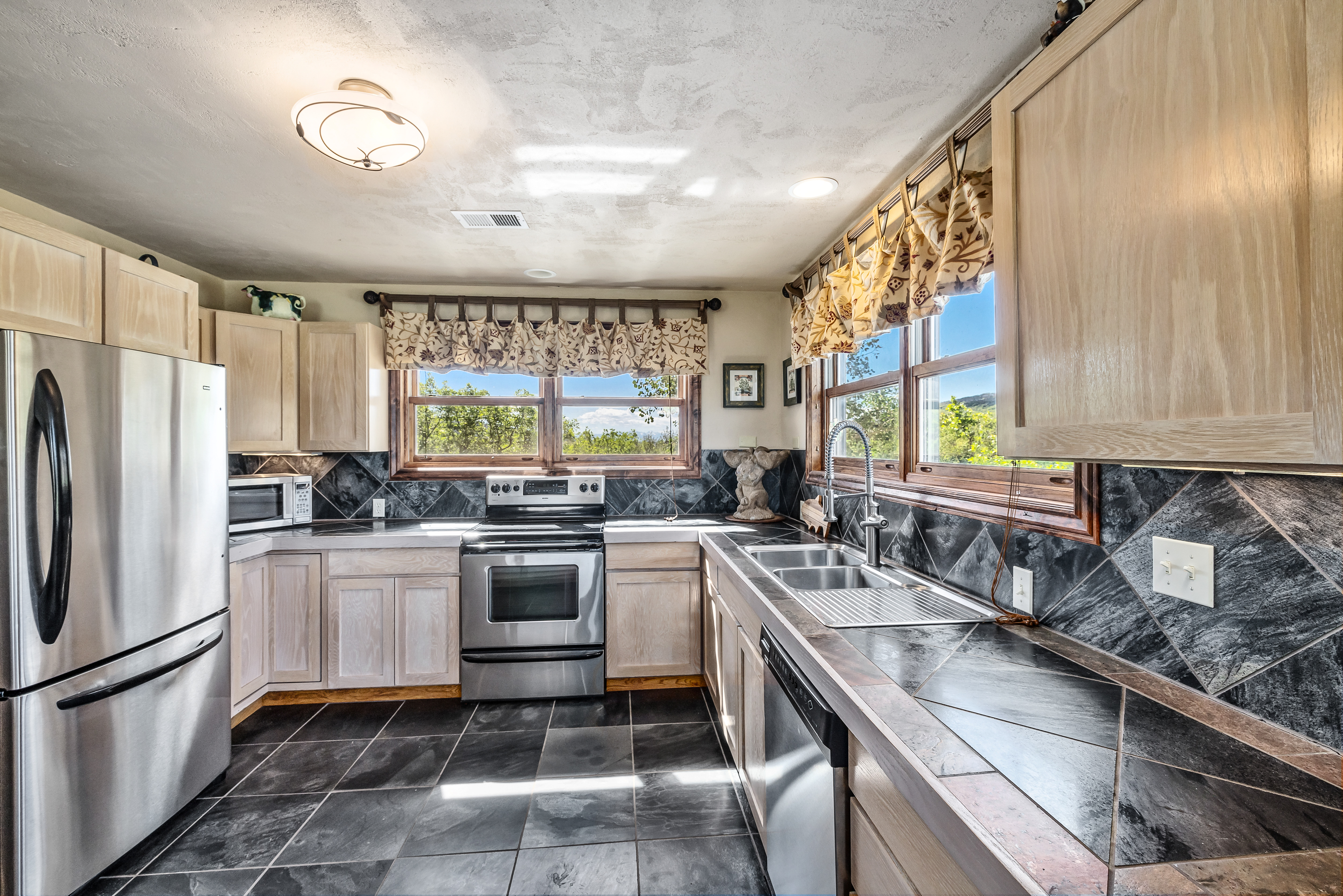 This kitchen features a rustic aesthetic with light-toned wooden cabinetry, dark slate tile flooring, and matching diagonal-patterned slate backsplashes. The space is equipped with stainless steel appliances, including a refrigerator, stove, and dishwasher, and is bathed in natural light from two large windows overlooking the outdoors. The overall impression is a cozy, functional workspace with a warm, earthy color palette.