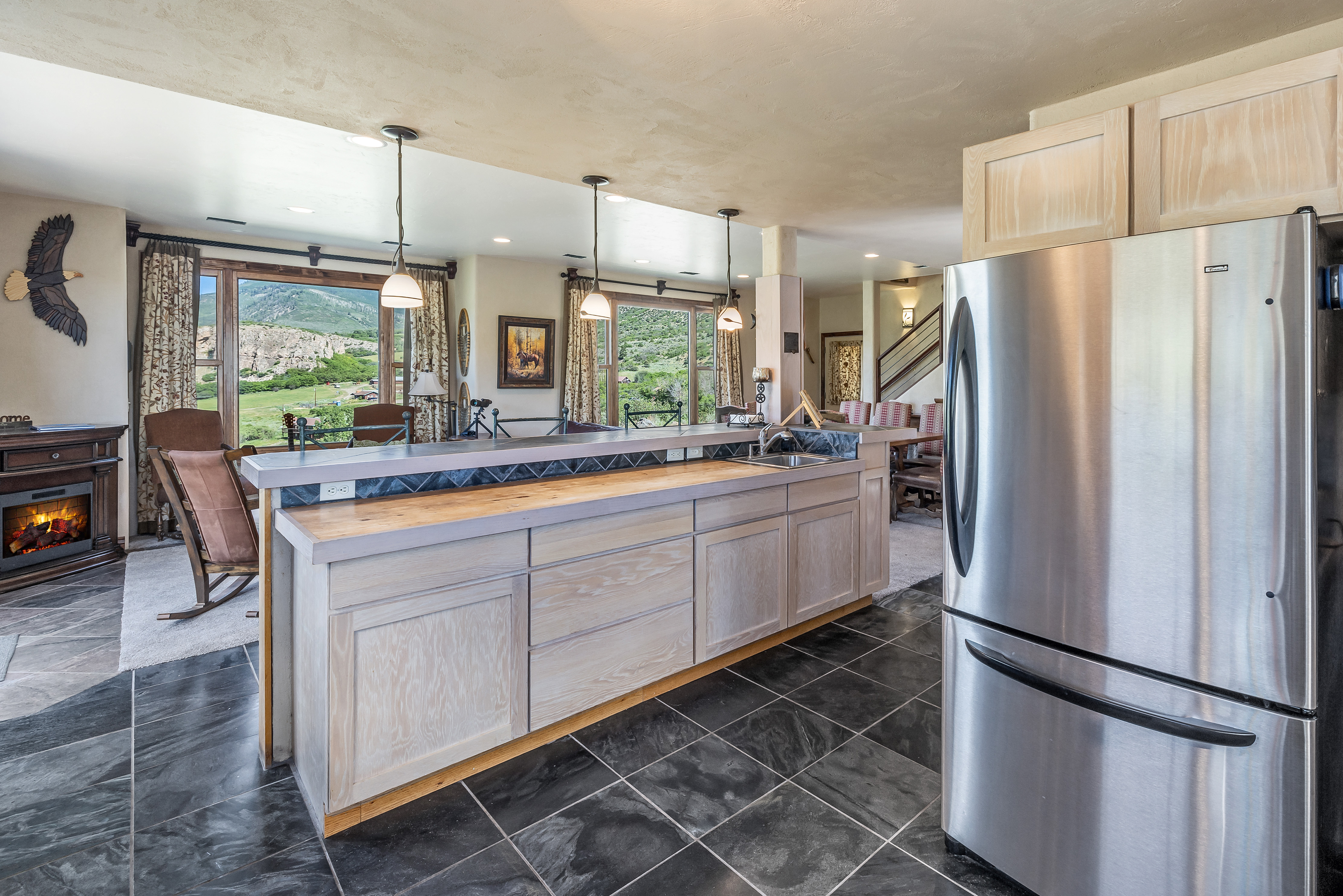 This spacious, open-concept kitchen features light-toned wooden cabinetry, a large island with a stone-tiled backsplash, and sleek stainless steel appliances. The room is anchored by dark, polished tile flooring and offers a seamless transition to a cozy living area with a fireplace and expansive mountain views through large windows. The warm, rustic-modern aesthetic creates an inviting atmosphere, perfectly blending indoor comfort with the natural beauty of the surrounding landscape.