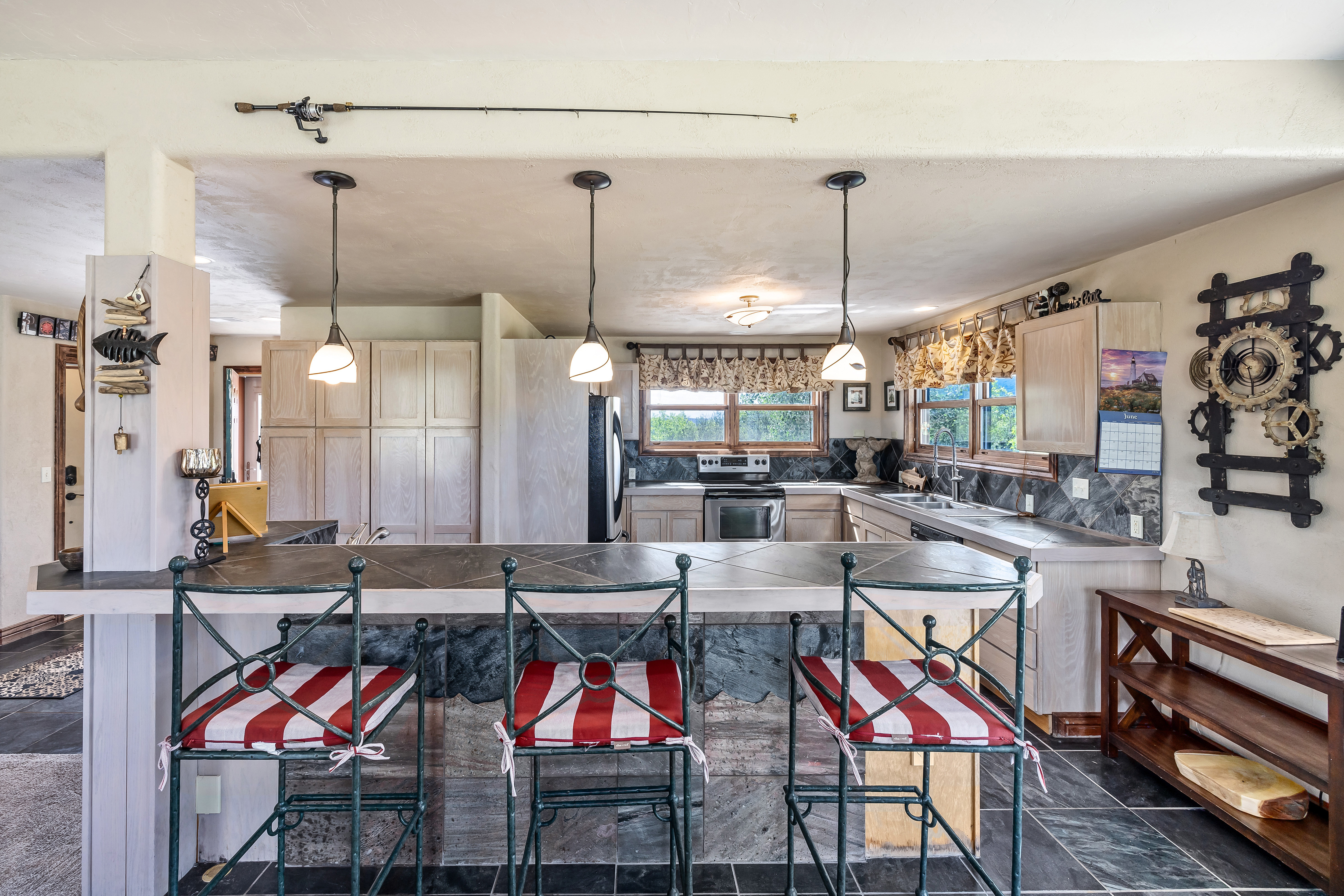 This rustic-themed kitchen features a spacious island with a dark stone countertop and three bar stools with red and white striped cushions. The space is accented with unique decor, including a fishing rod mounted above the breakfast bar and a large gear-themed wall clock, creating a cozy, cabin-like atmosphere. The kitchen is well-equipped with light wood cabinetry, stainless steel appliances, and slate-tiled surfaces, offering a warm and inviting space for cooking and entertaining.