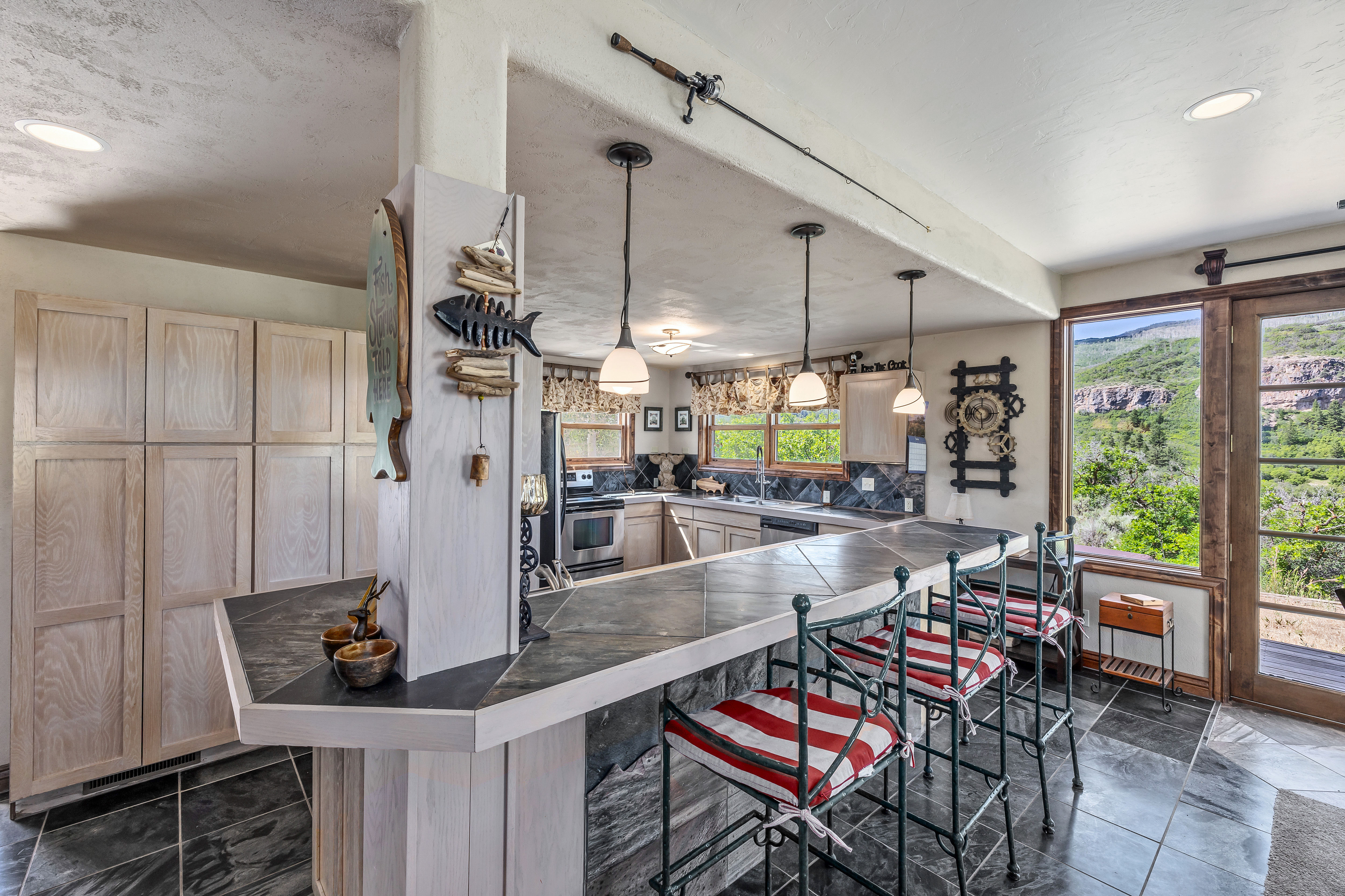 This rustic-style kitchen features a large, tiled breakfast bar with three red-and-white striped bar stools, serving as the central focal point. The space is accented with unique decor, including a fishing rod mounted on the ceiling beam and a fish-themed wall hanging on the support pillar. Large windows provide a scenic view of the surrounding landscape, creating a warm and inviting atmosphere.