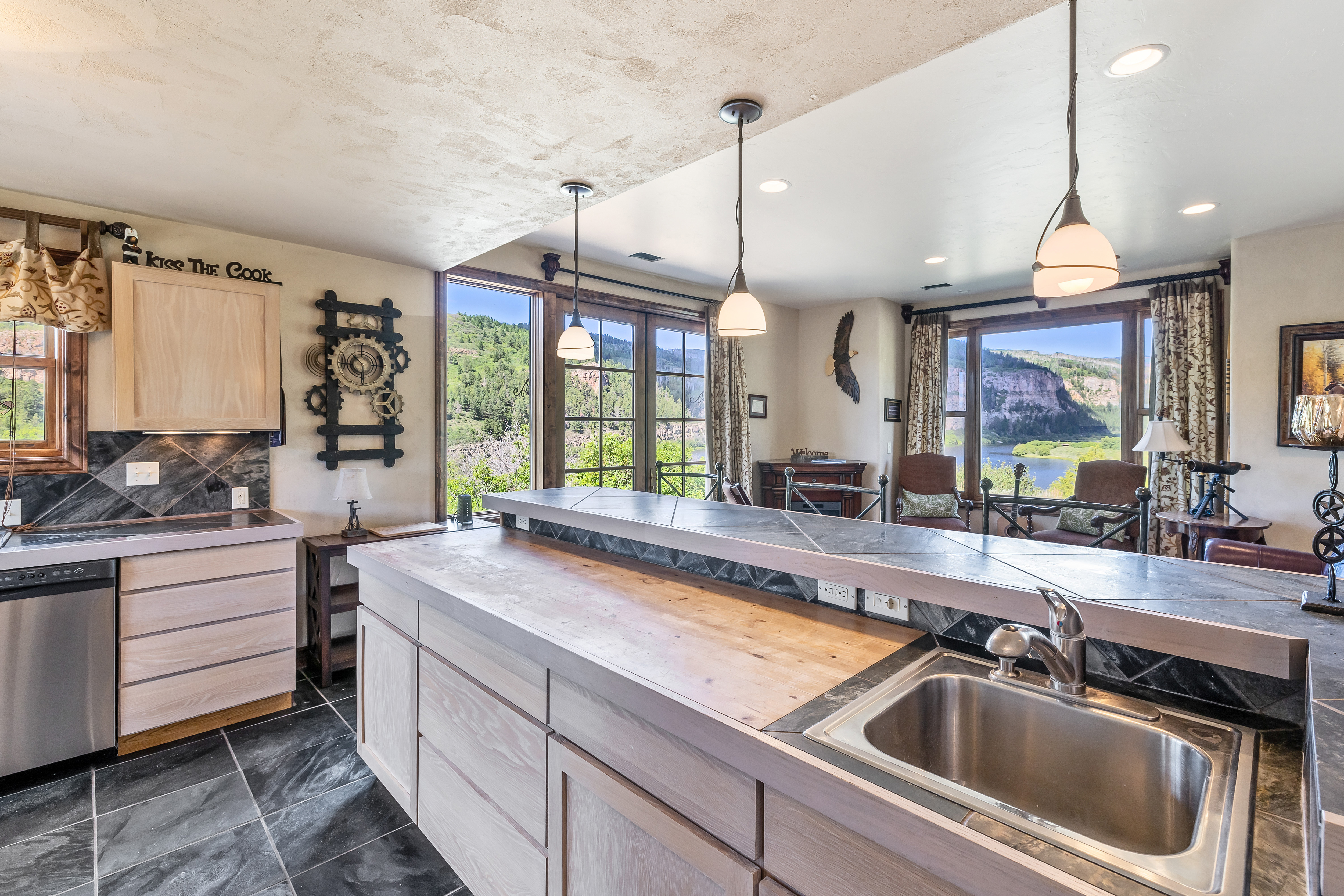 This rustic-style kitchen features a large island with a wooden countertop and a built-in stainless steel sink, complemented by light wood cabinetry and dark slate tile flooring. The space is illuminated by three pendant lights and offers expansive views of a scenic river and mountain landscape through large windows. The overall atmosphere is warm and inviting, blending natural textures with a functional layout perfect for entertaining.