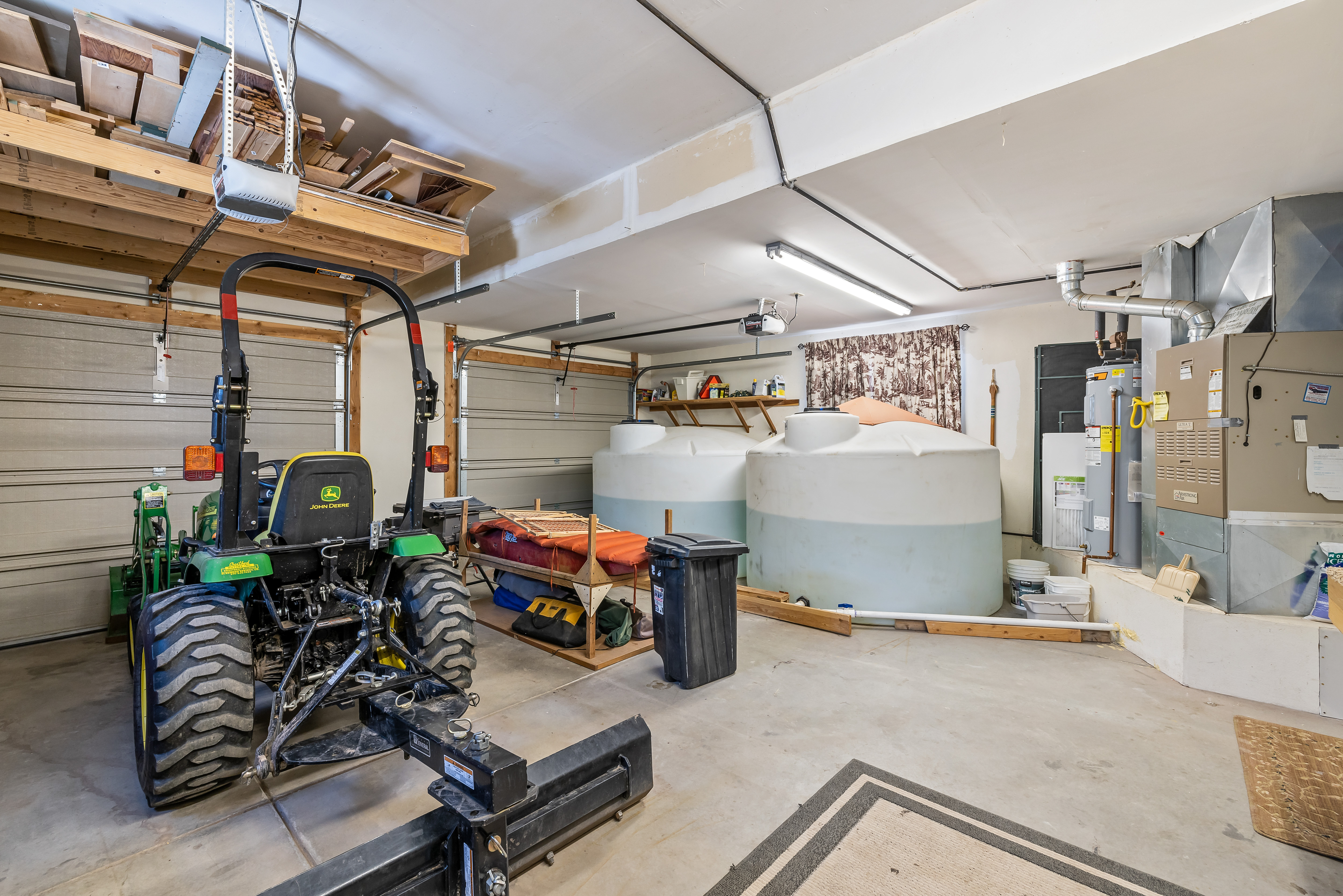 This spacious garage interior features a concrete floor and serves as a multi-functional utility and storage area. It houses a John Deere tractor, large water storage tanks, and a furnace system, with overhead wooden shelving providing ample storage space. The room is illuminated by a long fluorescent light fixture, highlighting the practical and utilitarian nature of the space.