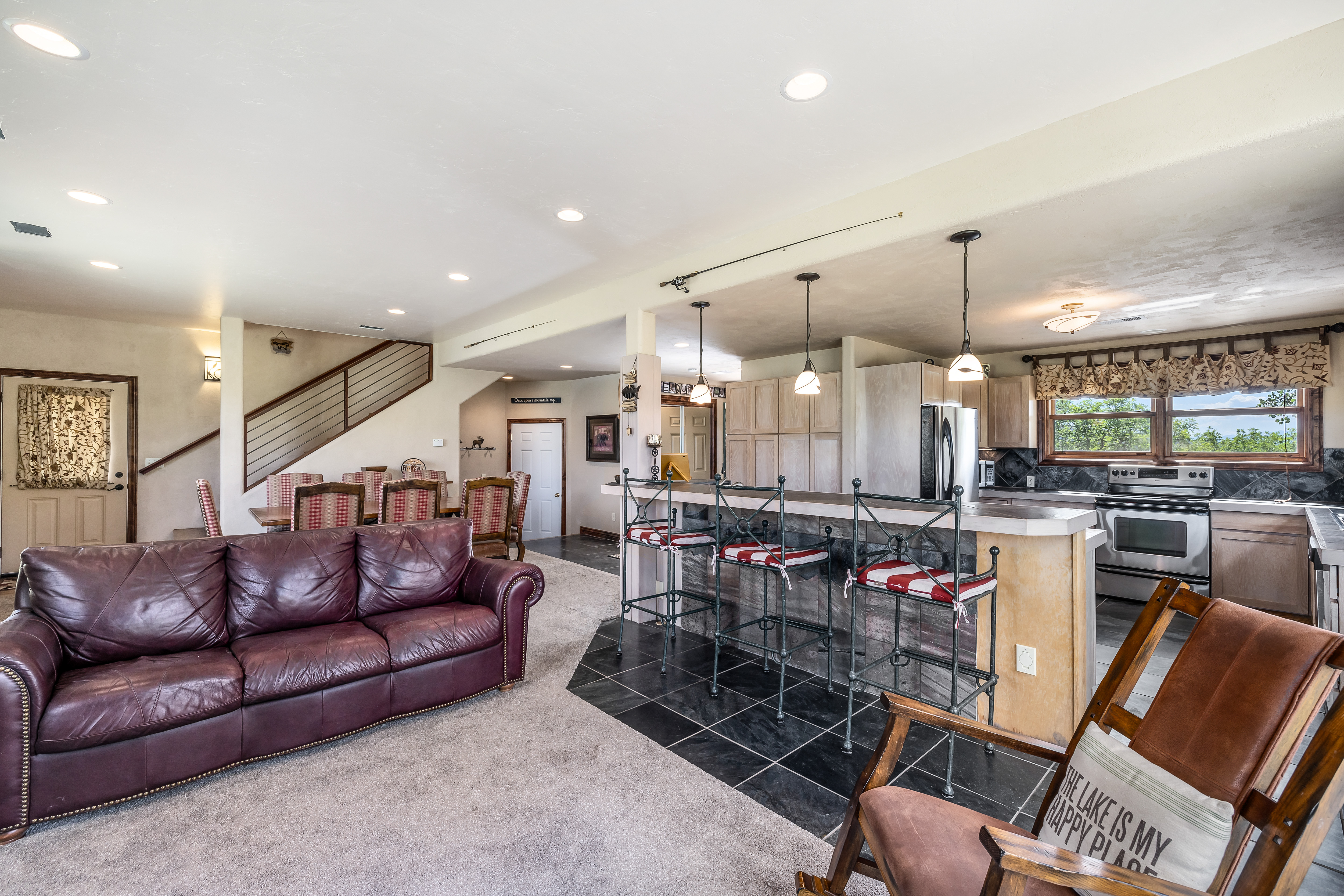 This open-concept living space features a plush, dark leather sofa set against a neutral carpeted floor, transitioning into a kitchen area with black slate tile flooring. A breakfast bar with three metal-framed stools separates the living area from the kitchen, which is equipped with stainless steel appliances and light wood cabinetry. The room is illuminated by recessed lighting and three pendant lights, with a staircase visible in the background, creating a warm and inviting atmosphere.