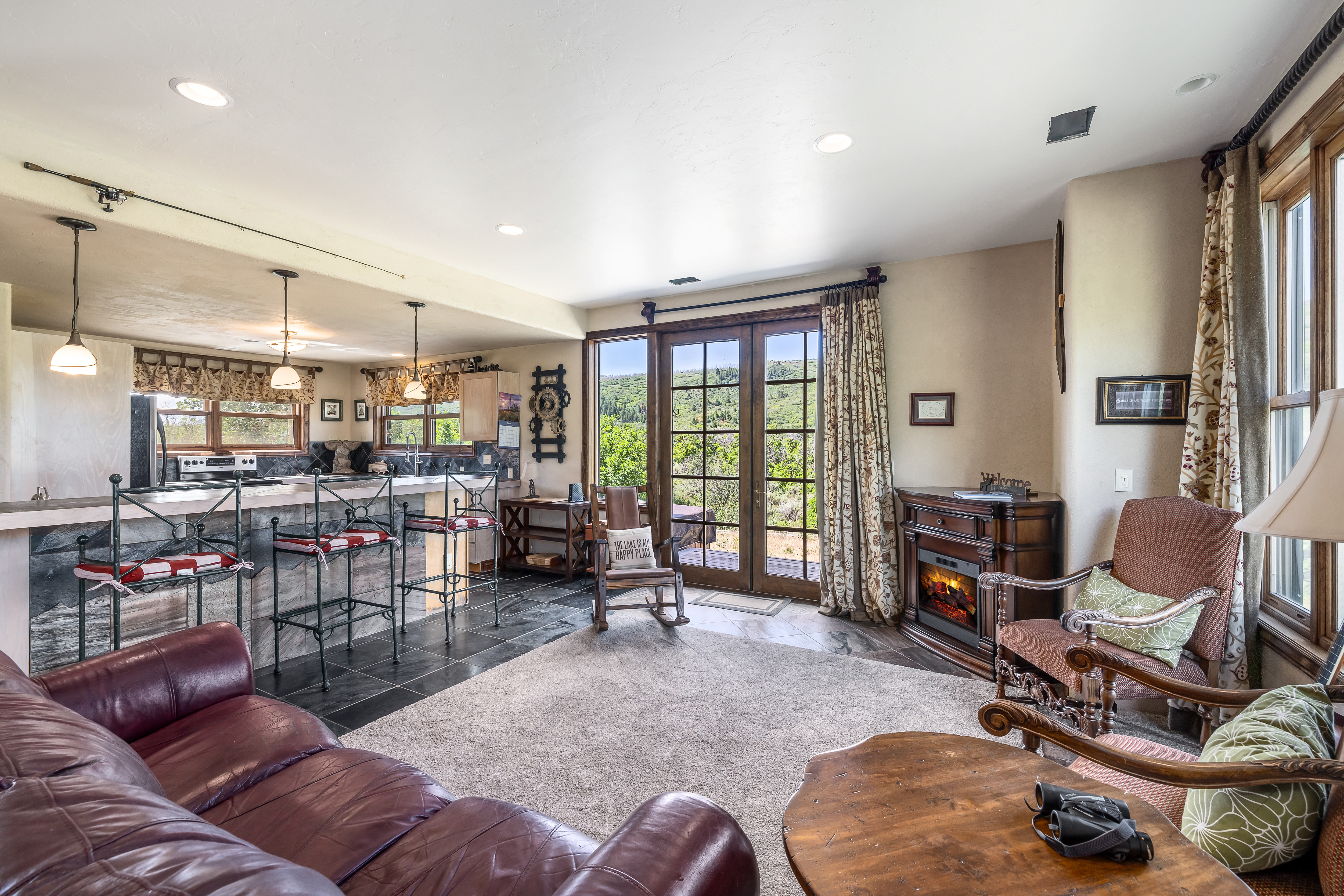 This open-concept living space seamlessly blends a cozy lounge area with a rustic kitchen, featuring a dark leather sofa, a stone-faced breakfast bar, and a warm electric fireplace. Large glass doors provide a picturesque view of the surrounding hills, while a rocking chair and patterned armchairs create a welcoming, cabin-inspired atmosphere. The room is bathed in natural light, highlighting the blend of tile and carpet flooring and the comfortable, lived-in aesthetic.