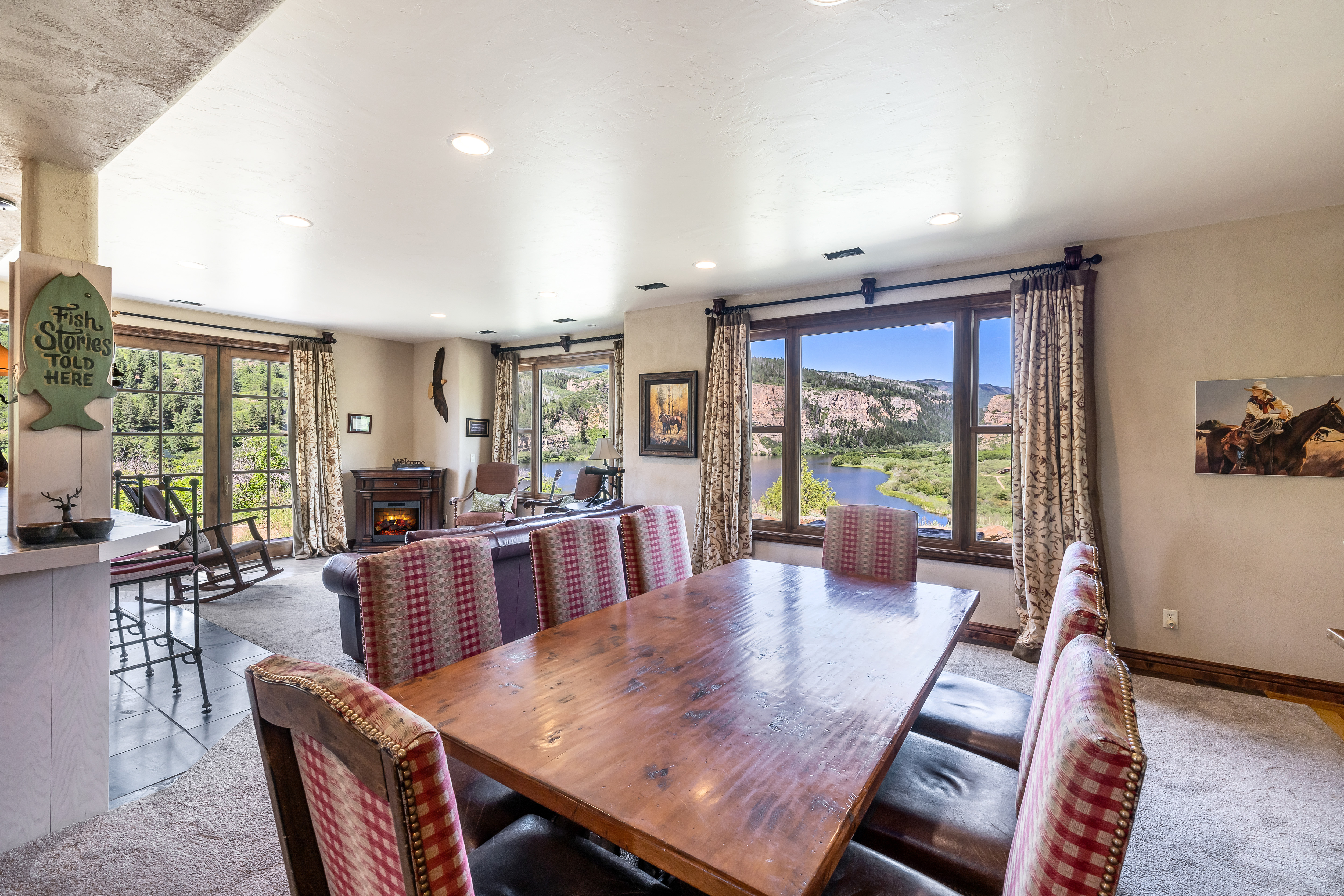 This inviting dining area features a large, rustic wooden table surrounded by chairs upholstered in a classic red and white checkered pattern. The room flows seamlessly into an adjacent living space, with expansive windows offering a breathtaking, panoramic view of a river winding through a scenic canyon landscape. The warm, earthy tones and natural light create a cozy, lodge-like atmosphere perfect for enjoying the surrounding wilderness.