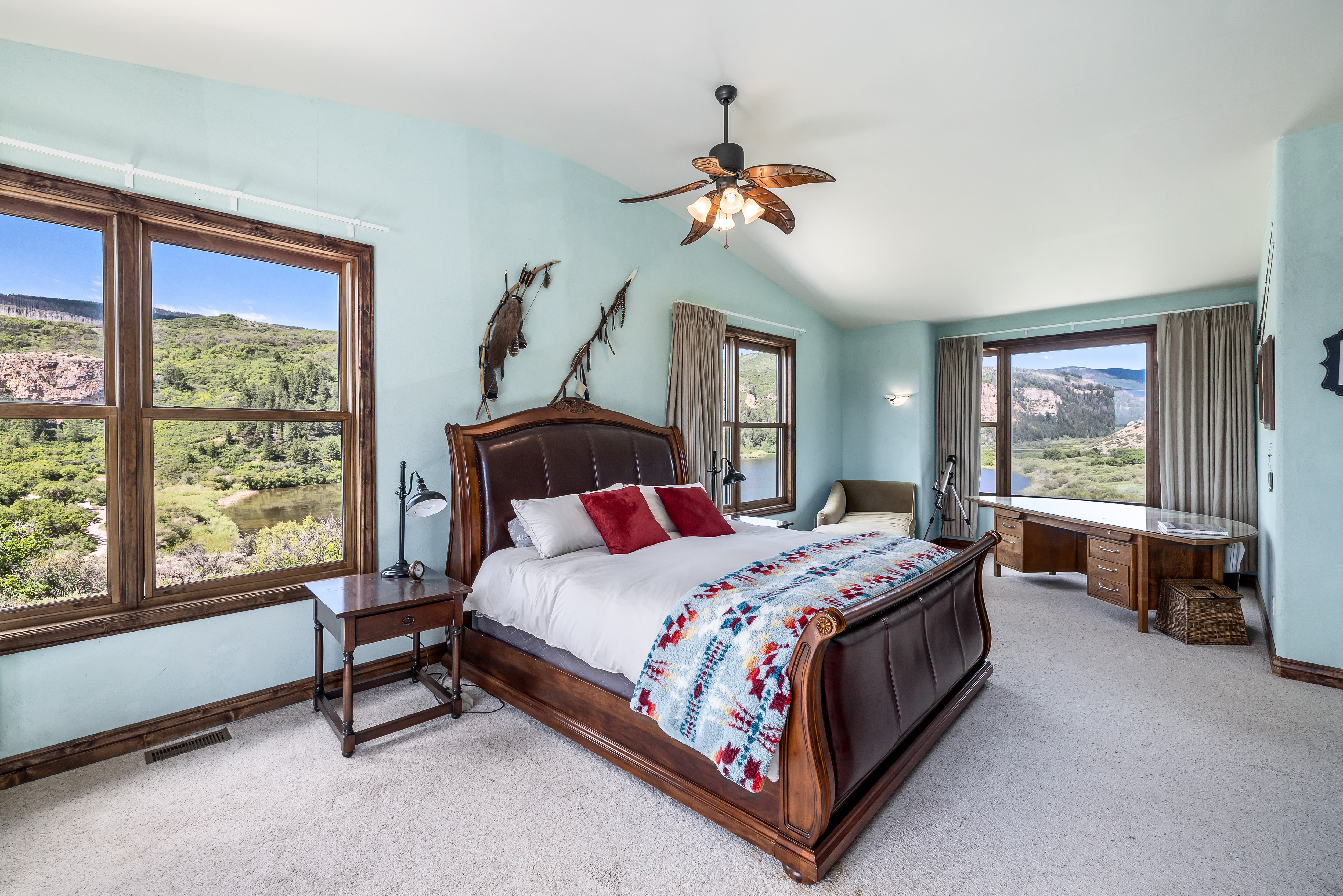 This spacious primary bedroom features a rich, dark wood sleigh bed as the centerpiece, complemented by a colorful patterned throw blanket and red accent pillows. The room is bathed in natural light from large windows that offer breathtaking, panoramic views of the surrounding mountains and river. A built-in wooden desk area and light blue walls create a serene, rustic-inspired atmosphere perfect for relaxation.