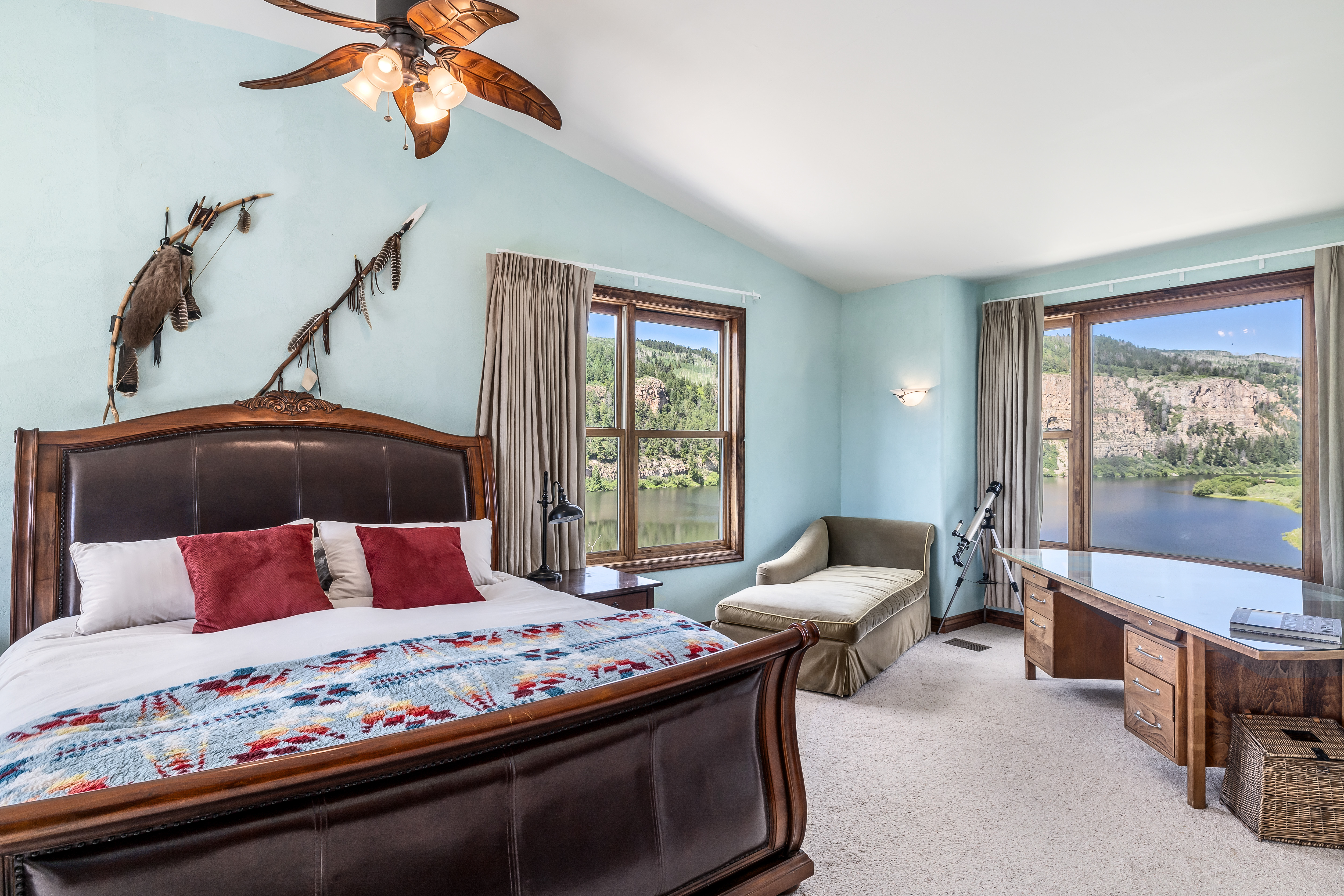 This spacious primary bedroom features a large leather-framed bed with a colorful patterned quilt and red accent pillows, set against light blue walls adorned with rustic wall art. The room includes a comfortable chaise lounge and a wooden desk positioned to take advantage of the stunning panoramic views of the lake and mountains through large windows. A unique palm-leaf ceiling fan adds a touch of tropical flair to the serene, nature-inspired retreat.